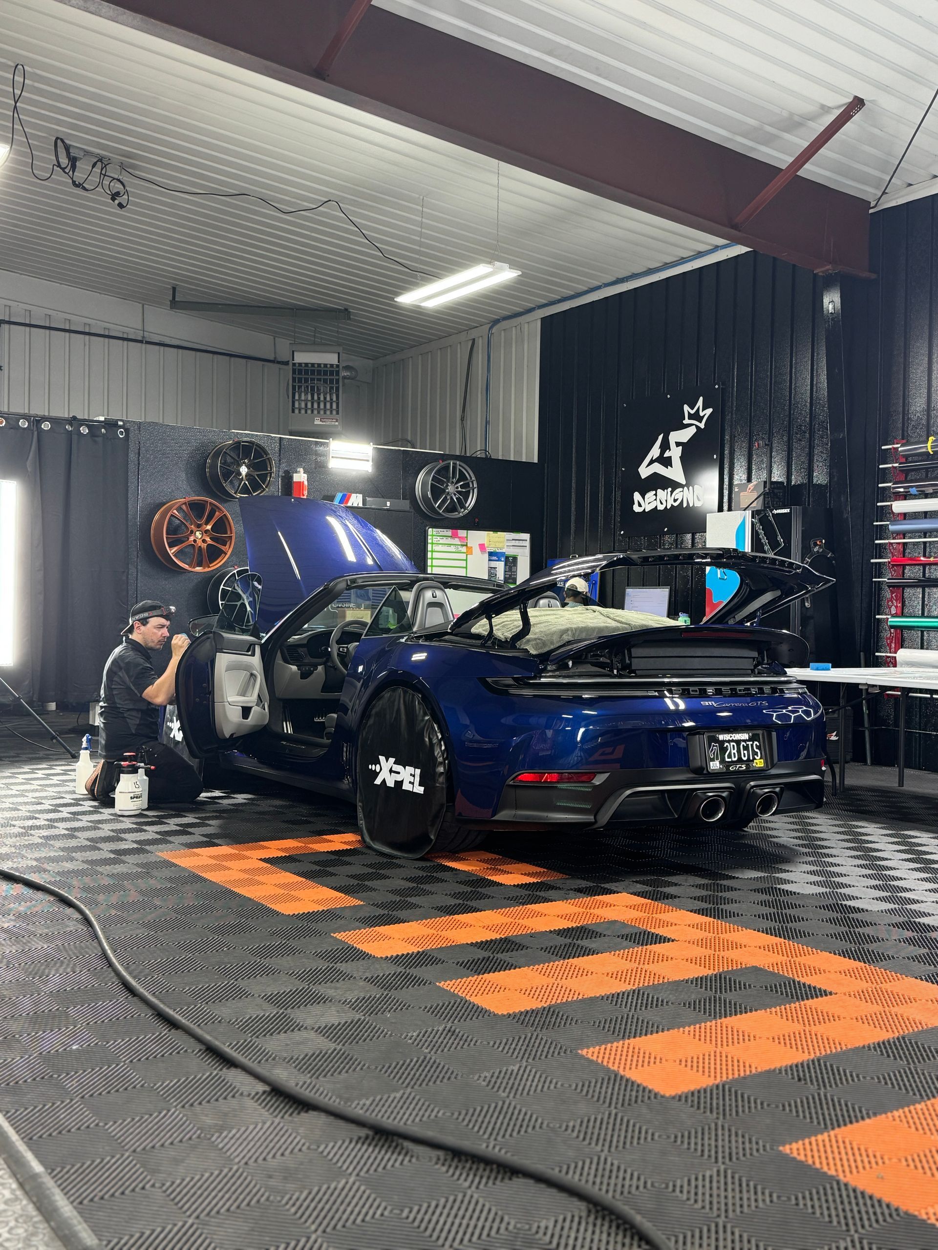 Mechanic working on a blue convertible sports car inside a garage with black and orange flooring.