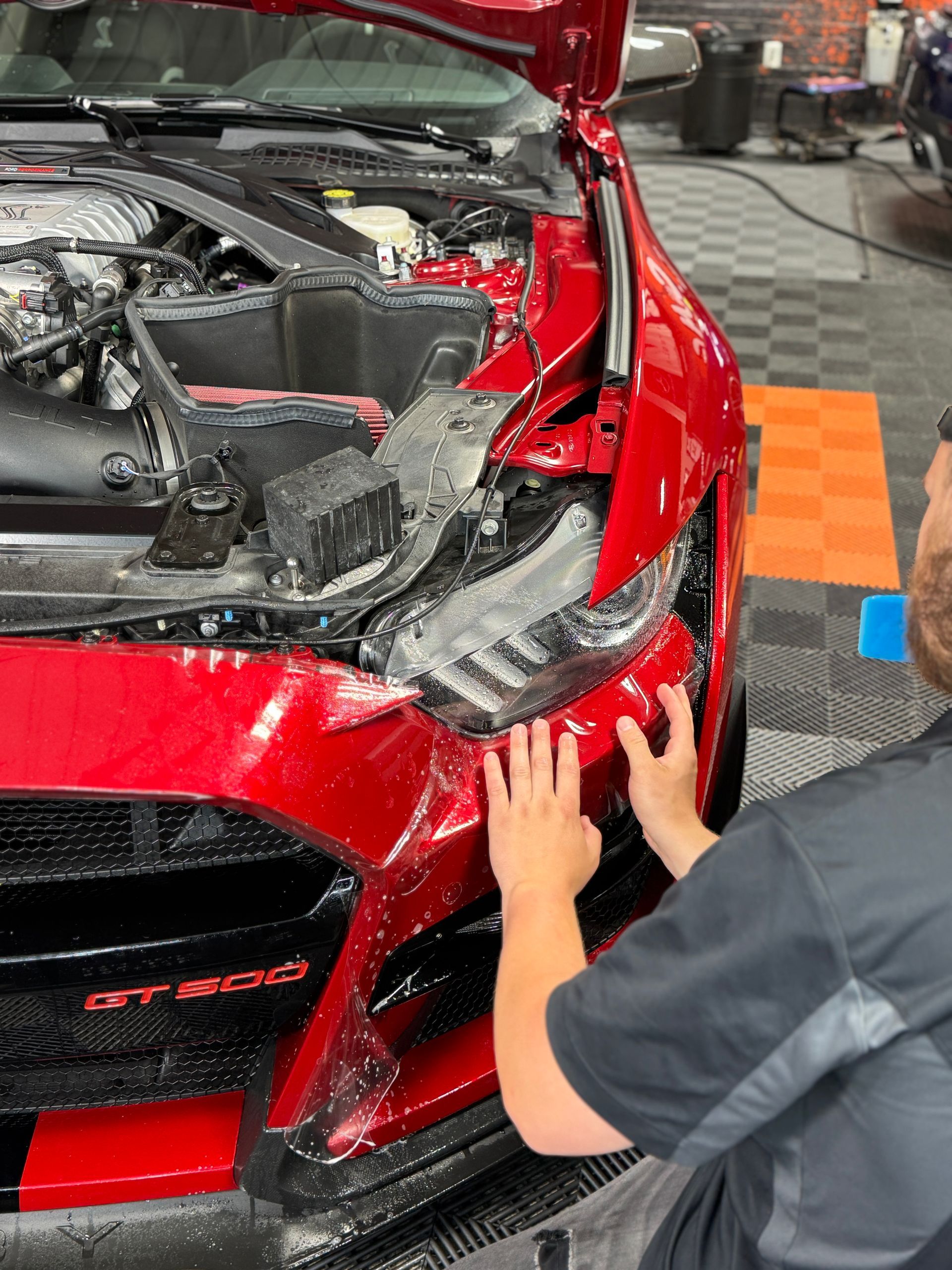 Person applying protective film to the front of a red Ford Mustang GT500 in a shop.