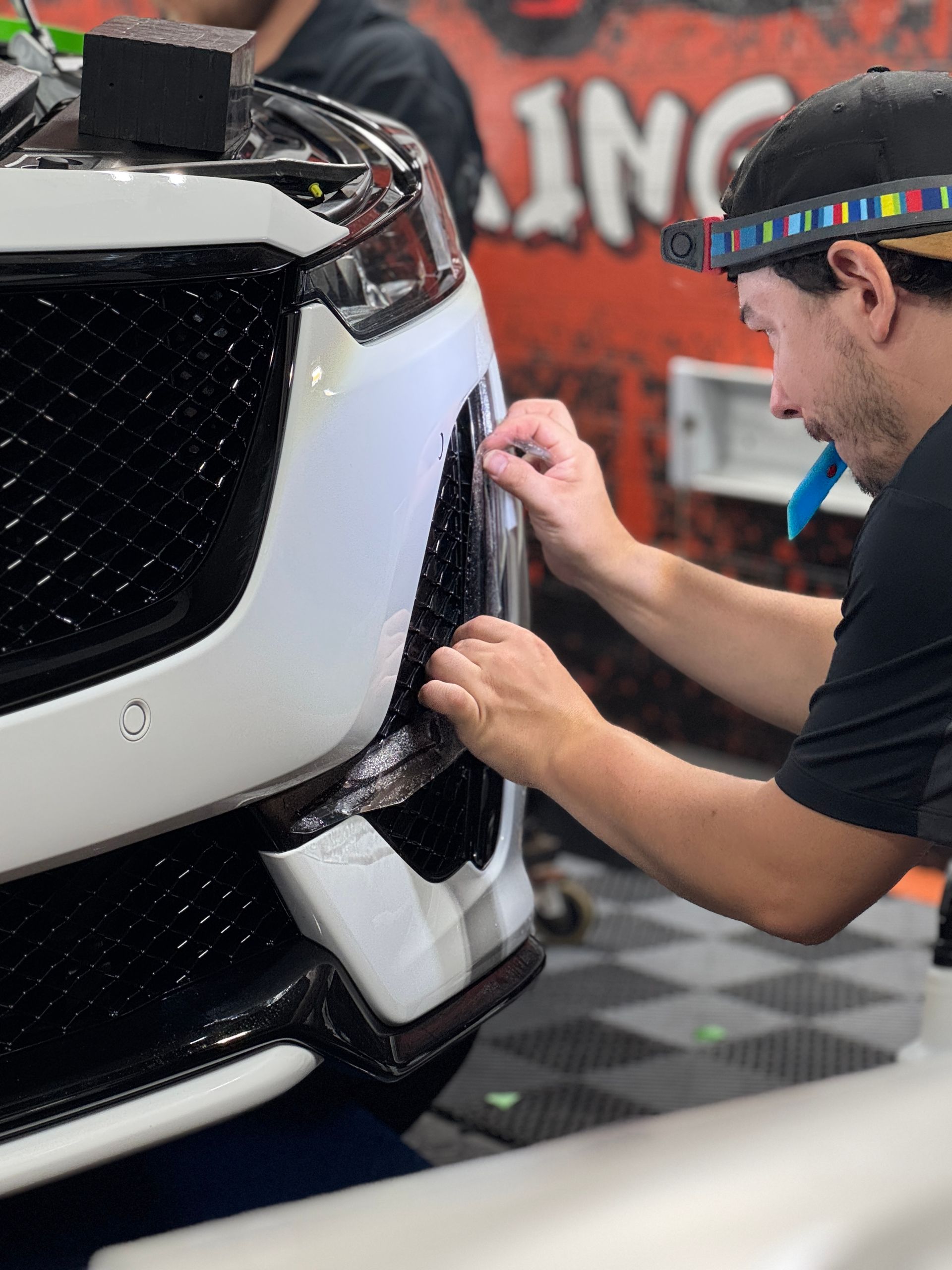Man applying a film to a car's black grille, in a shop with orange and black accents.