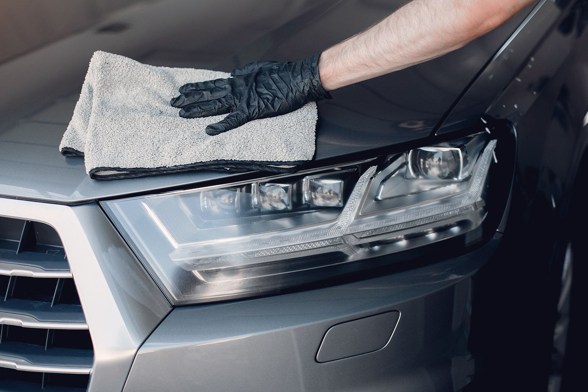 Gloved hand wiping a gray car hood with a gray microfiber cloth; close-up.