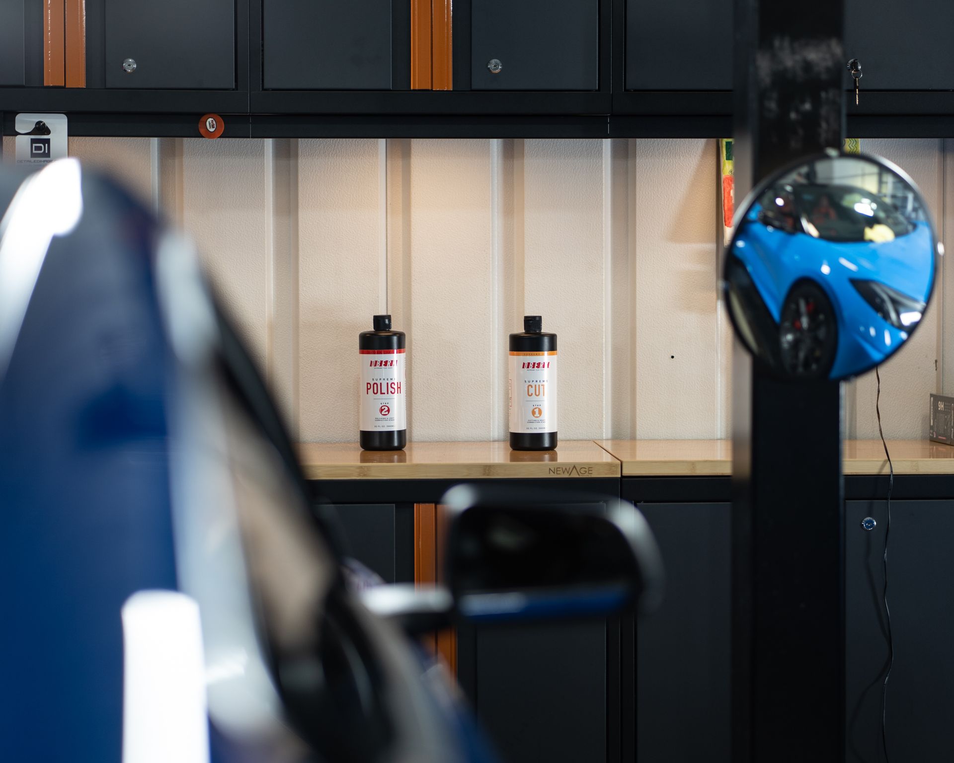 Blue car in a garage with bottles on a shelf. A round mirror reflects another blue car.