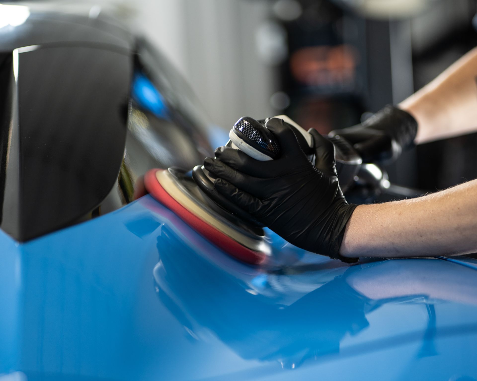 Hands wearing black gloves polishing blue car paint with a buffing machine.