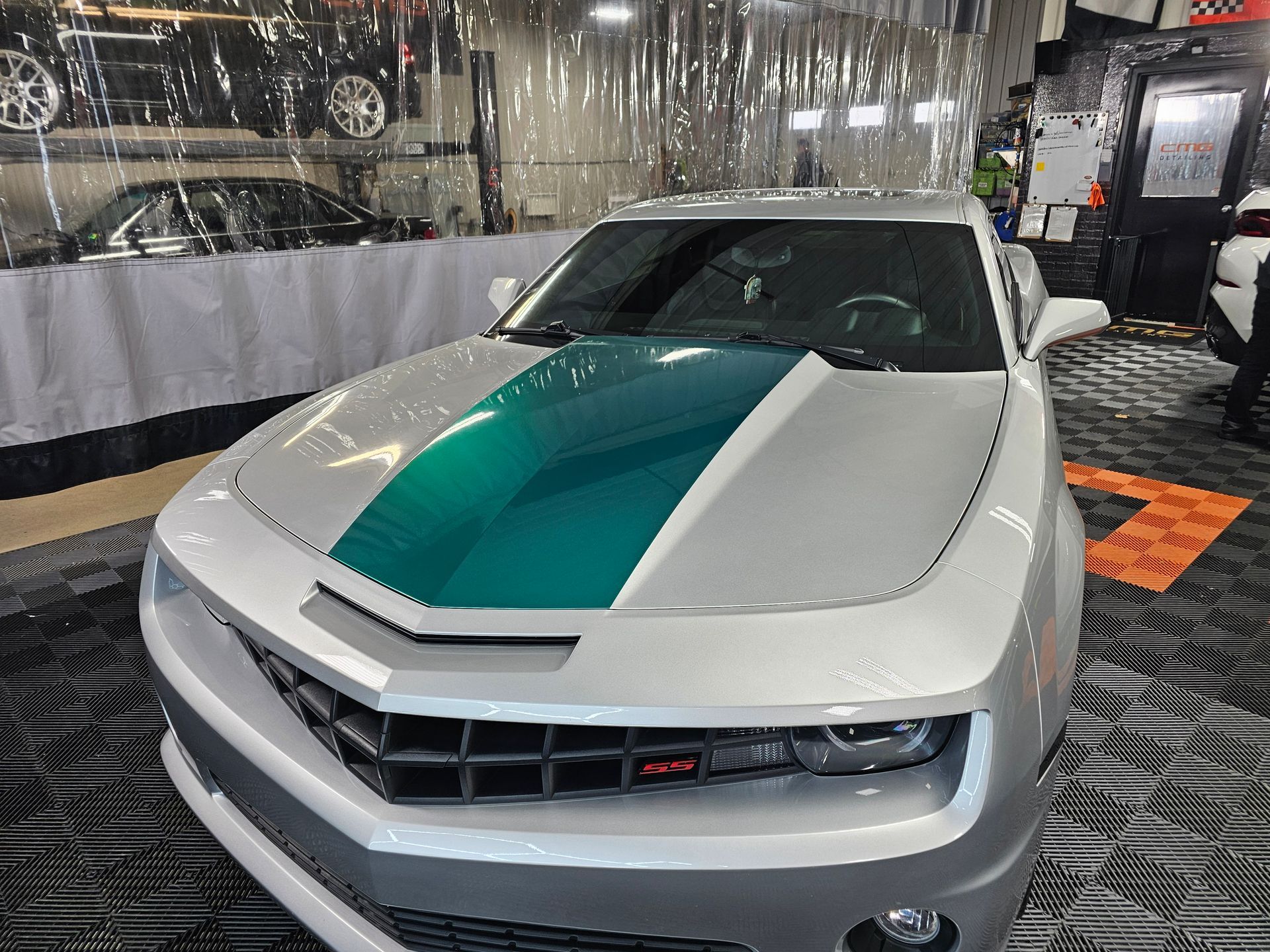 Silver Chevy Camaro with teal racing stripe parked in a garage.