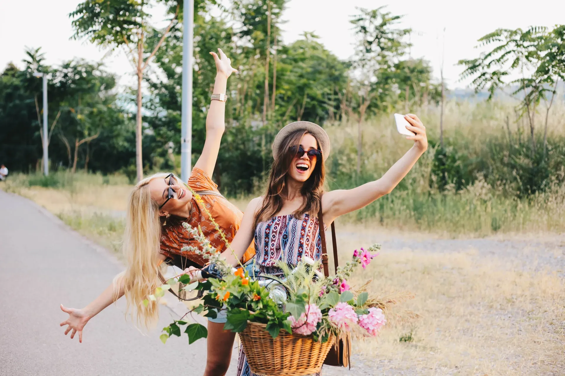 Two women take a playful selfie outdoors beside a flower-filled bicycle basket.
