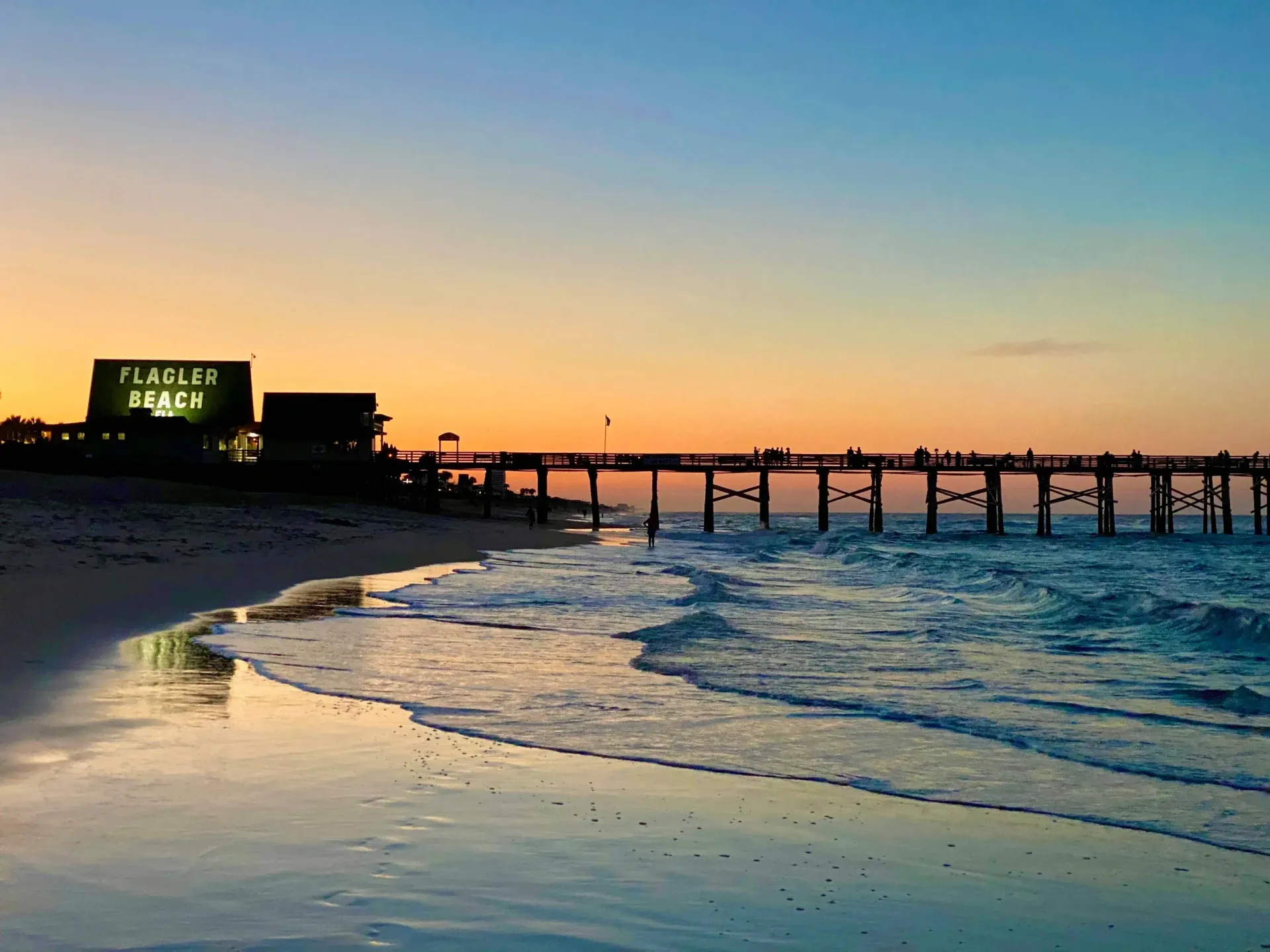 Sunset at the beach with a wooden pier and a Flagler Beach building; silhouetted people on the pier.