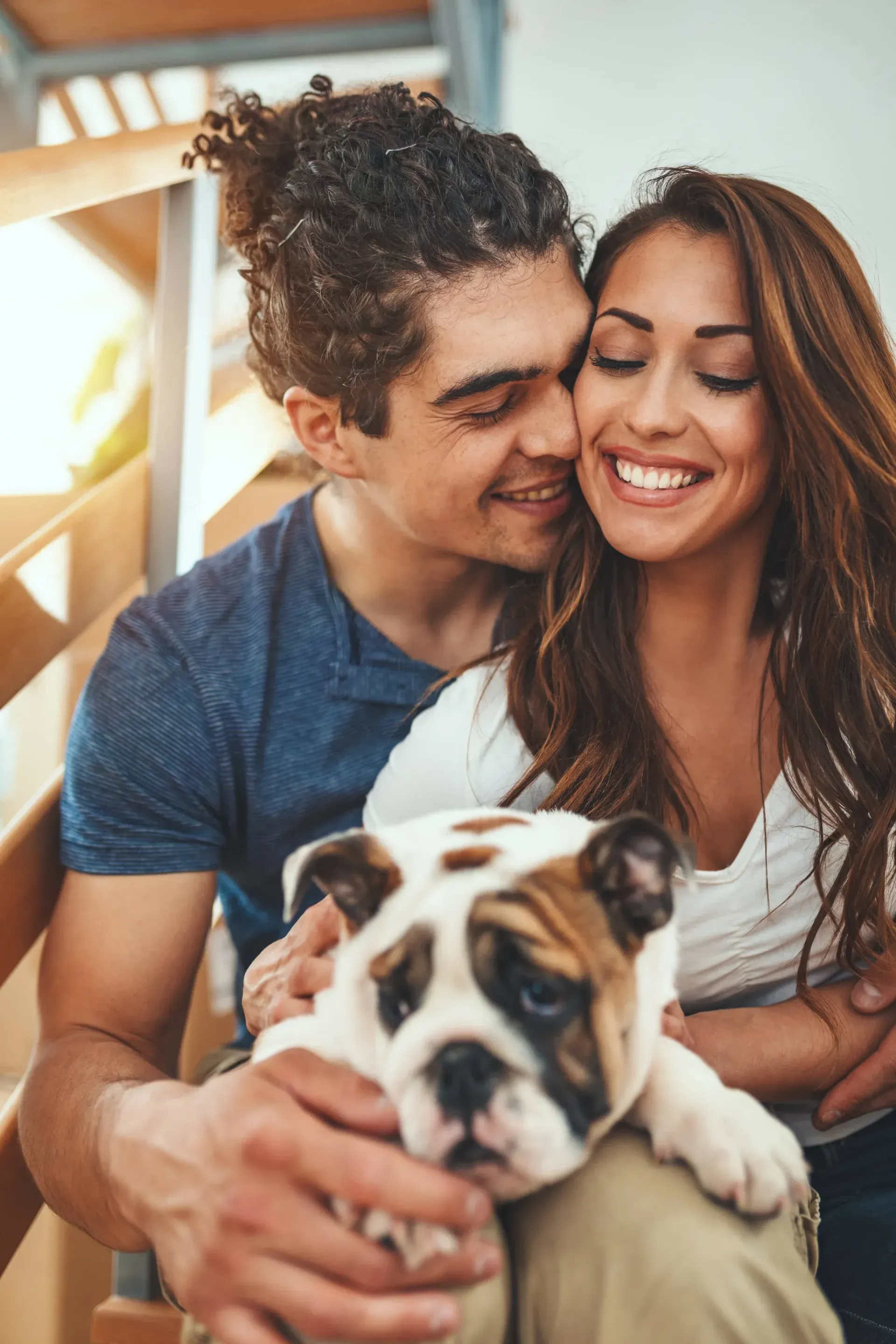 Smiling couple cuddling with a bulldog puppy on a balcony.