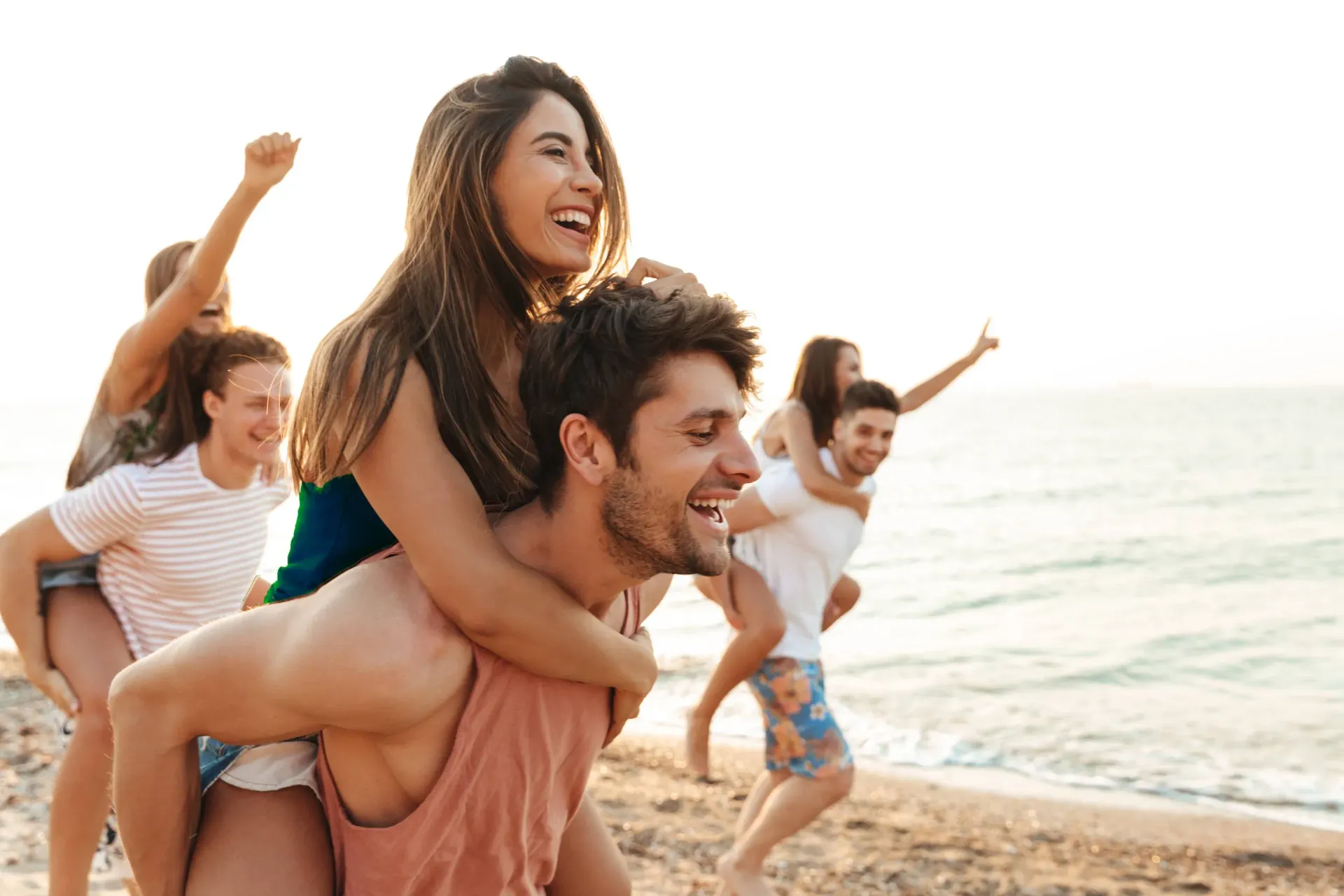 Group of friends at the beach; two people giving piggyback rides. Everyone is smiling and laughing.