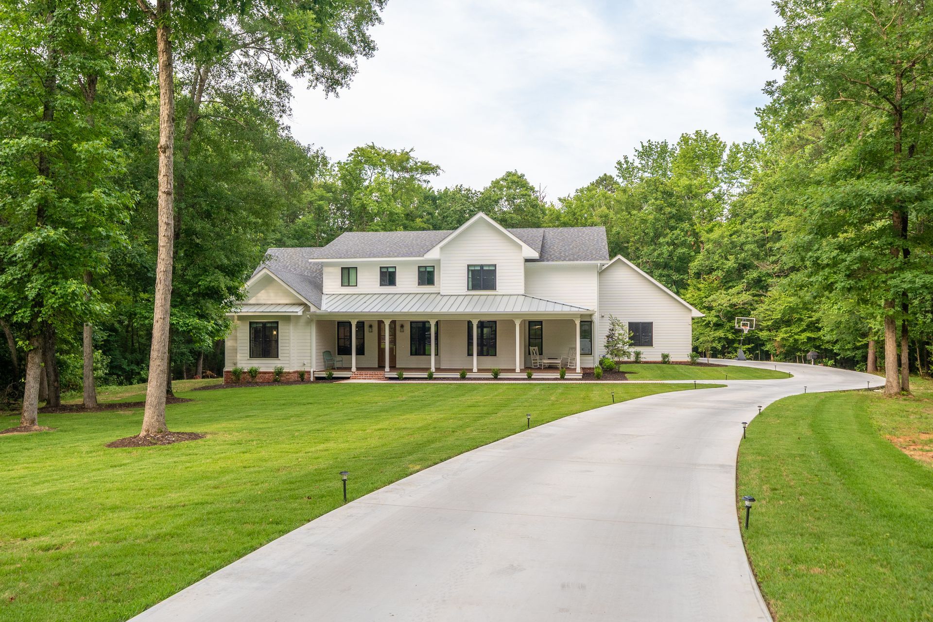 A white, two-story farmhouse with a metal roof and a long concrete driveway, surrounded by lush green trees and lawn.