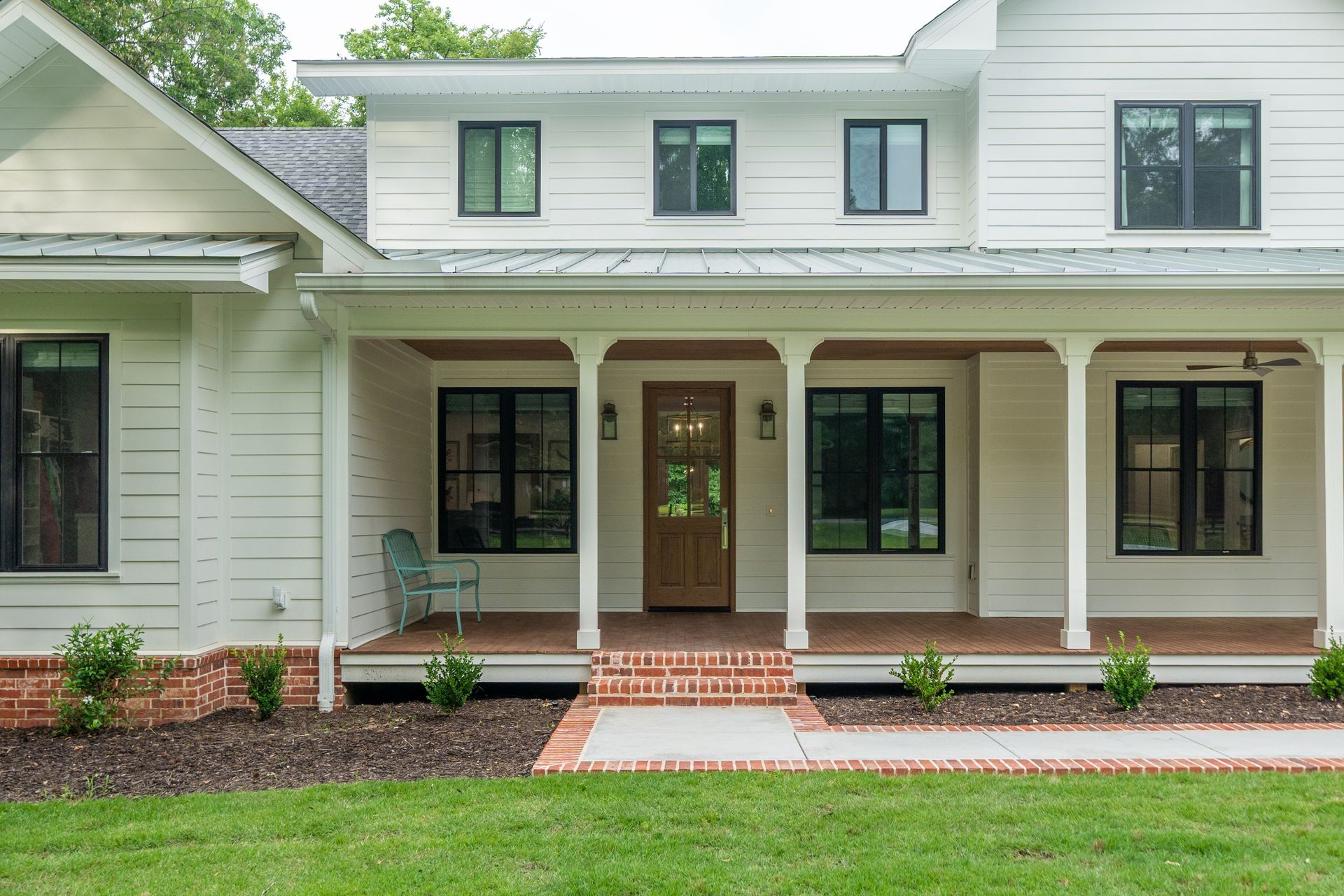 A modern white farmhouse with a large front porch, dark-framed windows, a wooden front door, and a brick base.