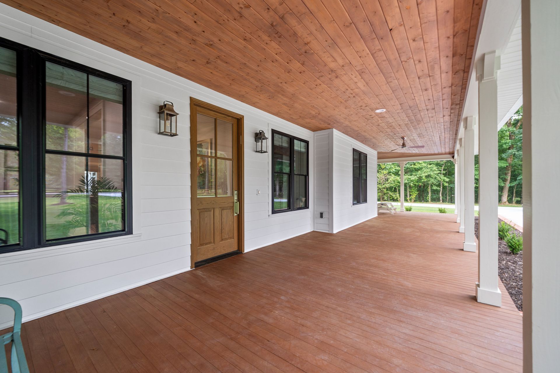 A wide covered porch with a wood-plank ceiling and floor, white brick walls, black-framed windows, and a wood front door.