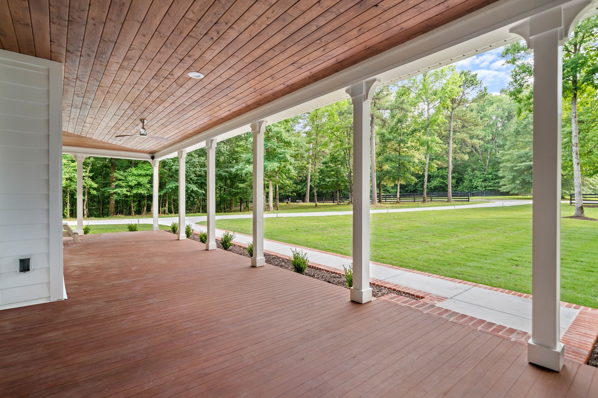 A spacious porch with a wood-paneled ceiling, red brick flooring, and white columns overlooking a grassy yard and trees.