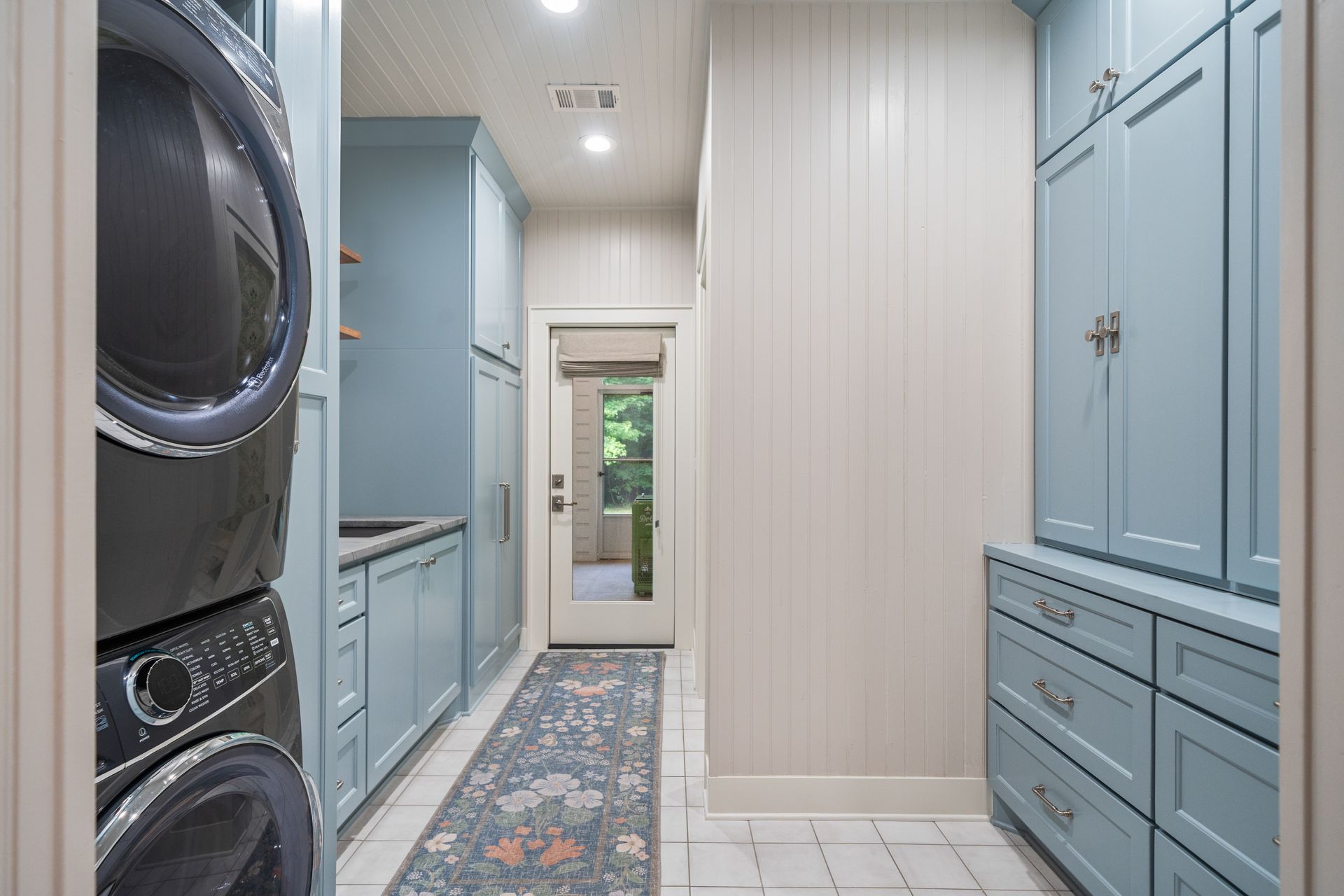 A laundry room with a stacked washer and dryer, light blue cabinetry, a patterned runner rug, and a glass-paneled door.