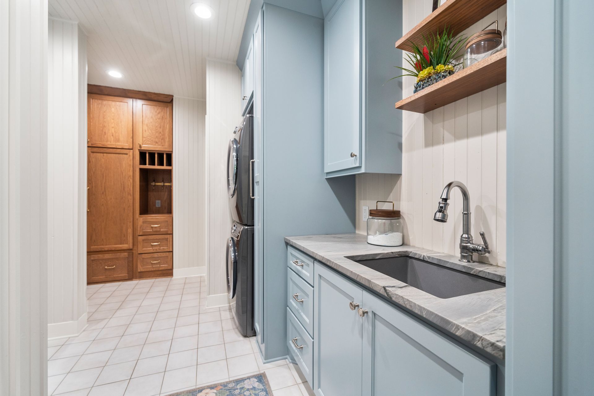 A laundry room with blue cabinets, a gray sink, light-colored tile floors, and a tall wooden storage unit in the back.