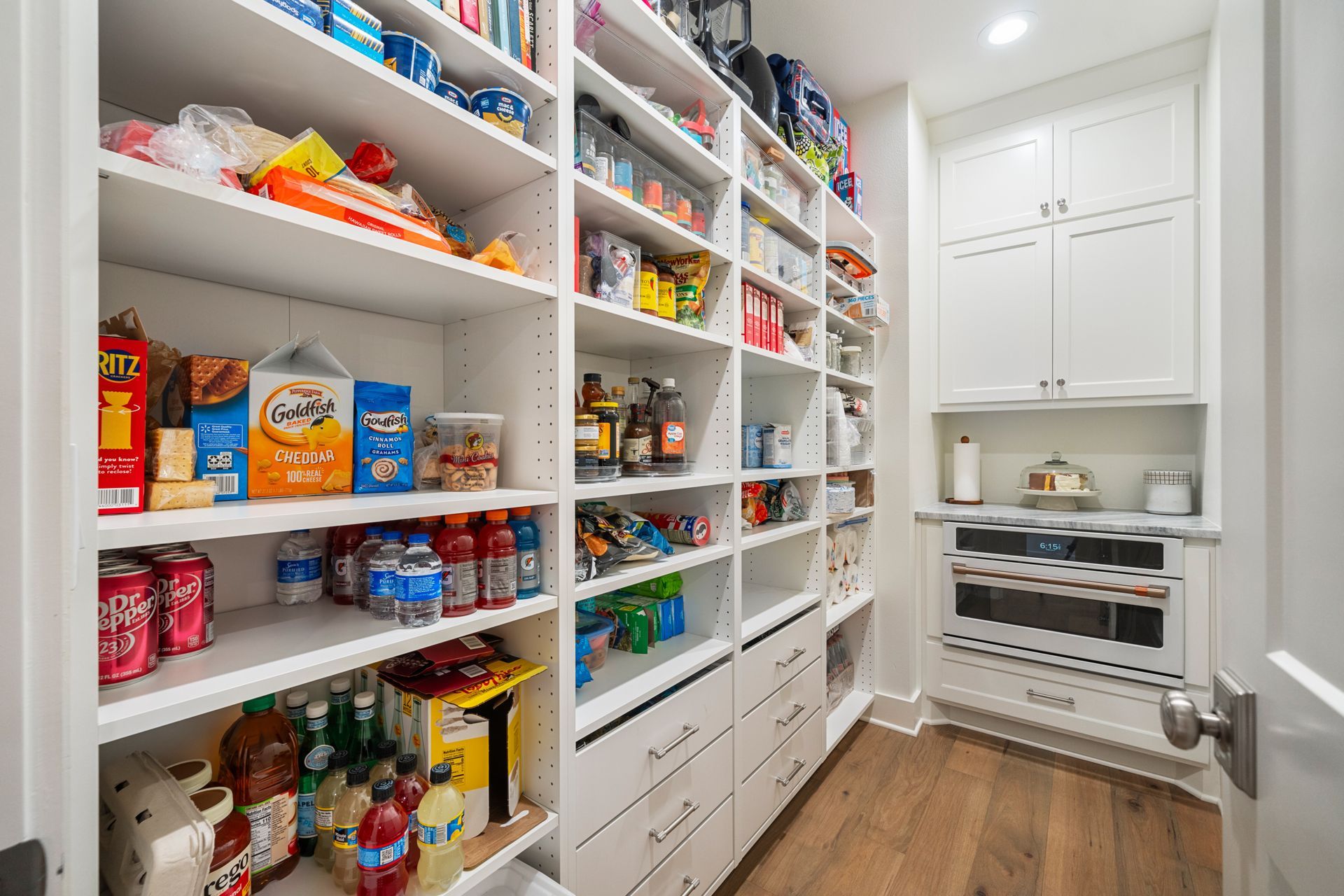 A well-organized white pantry with shelves stocked with food items, drawers, and a built-in microwave on a wood floor.