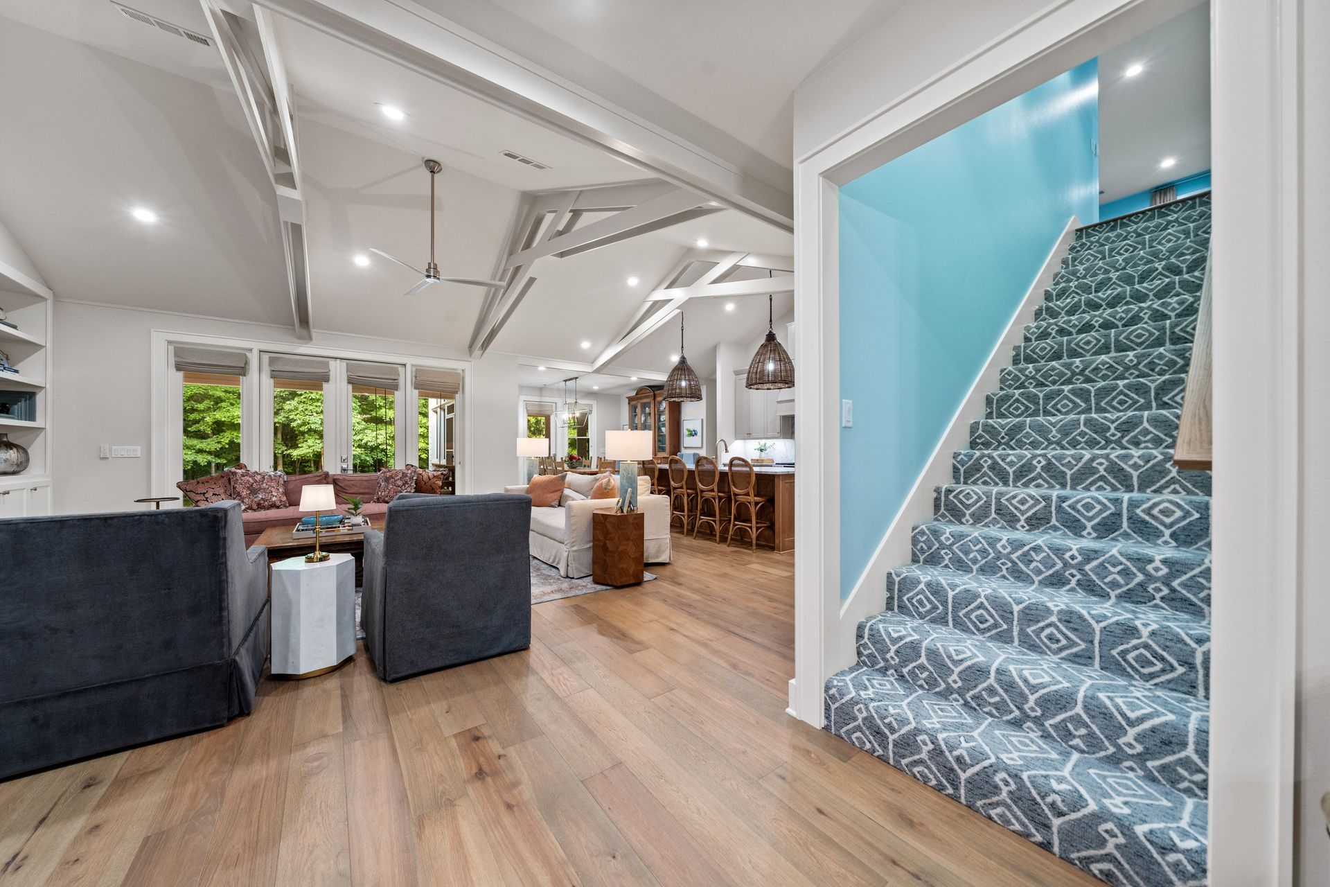 A spacious living area with hardwood floors, neutral furniture, and a blue-carpeted staircase leading to an upper level.