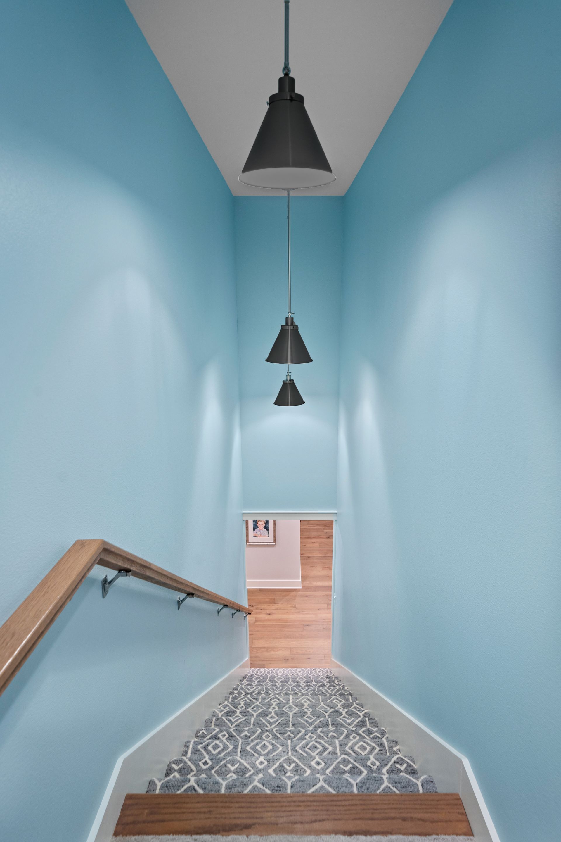 Staircase with light blue walls, gray patterned runner, wooden handrail, and three hanging black pendant lights.