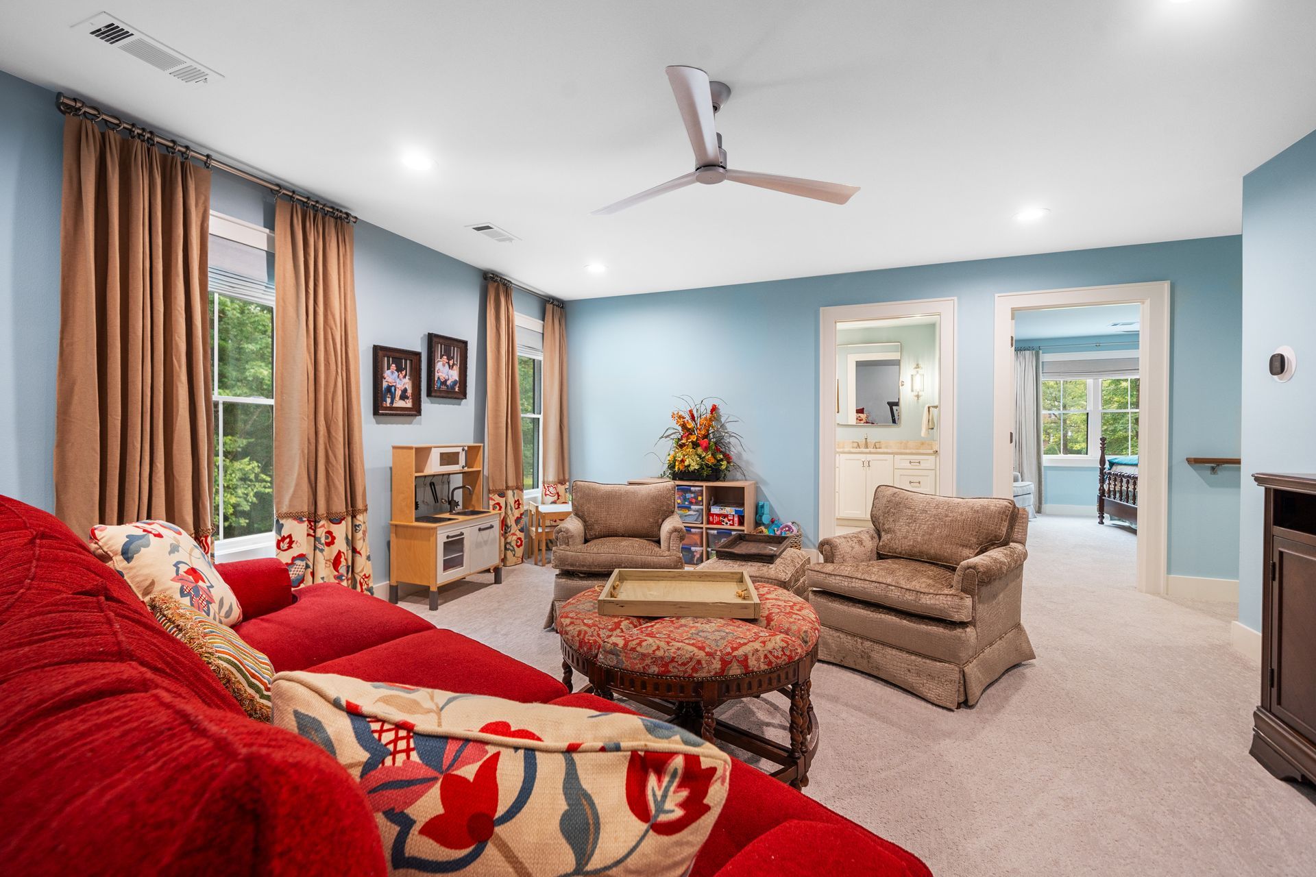 A bright living room with light blue walls, a red sofa, two beige armchairs, and floor-to-ceiling brown curtains.