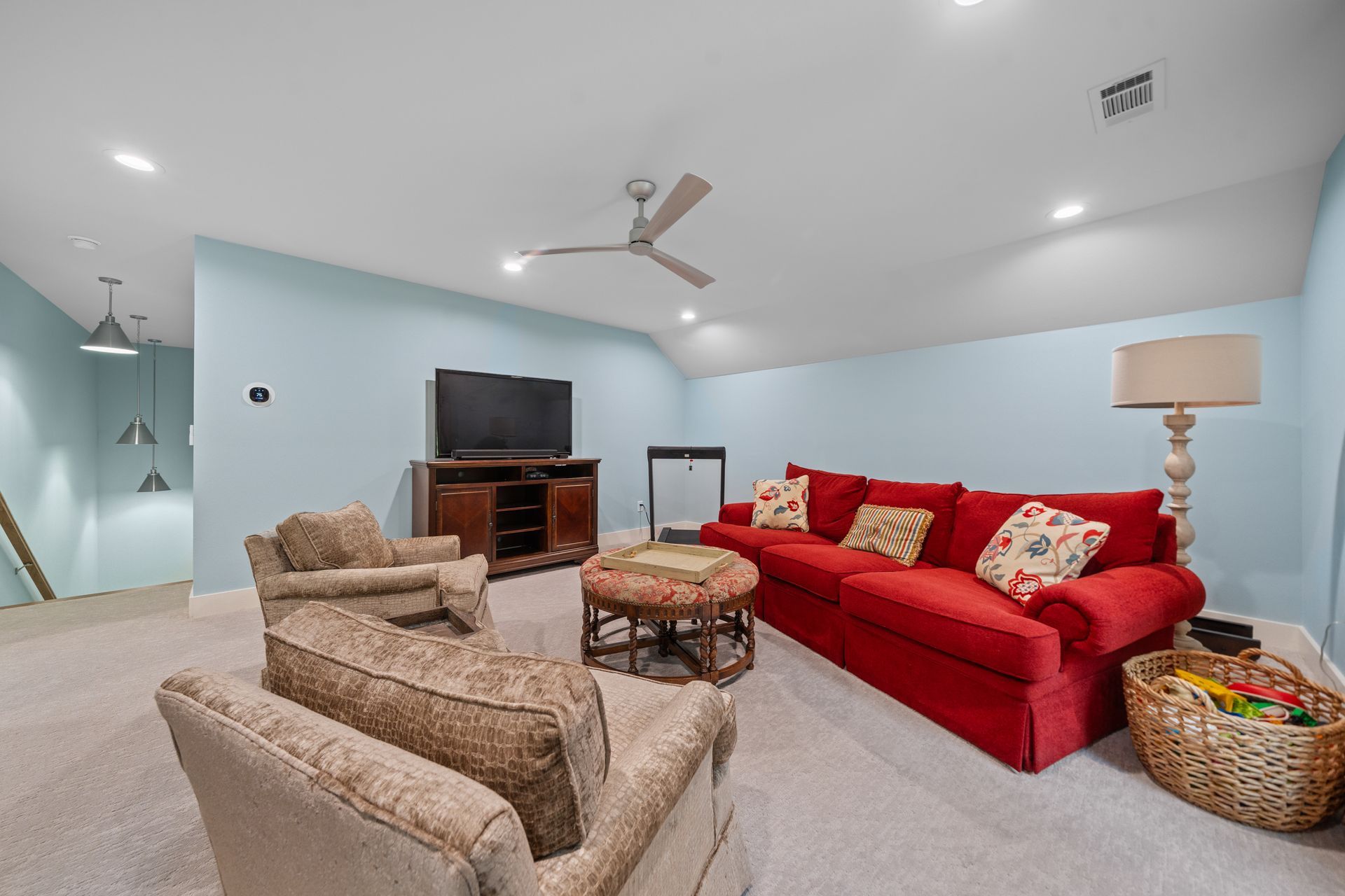 A cozy living room with a red sofa, two beige armchairs, a wooden media console, a round ottoman, and light blue walls.