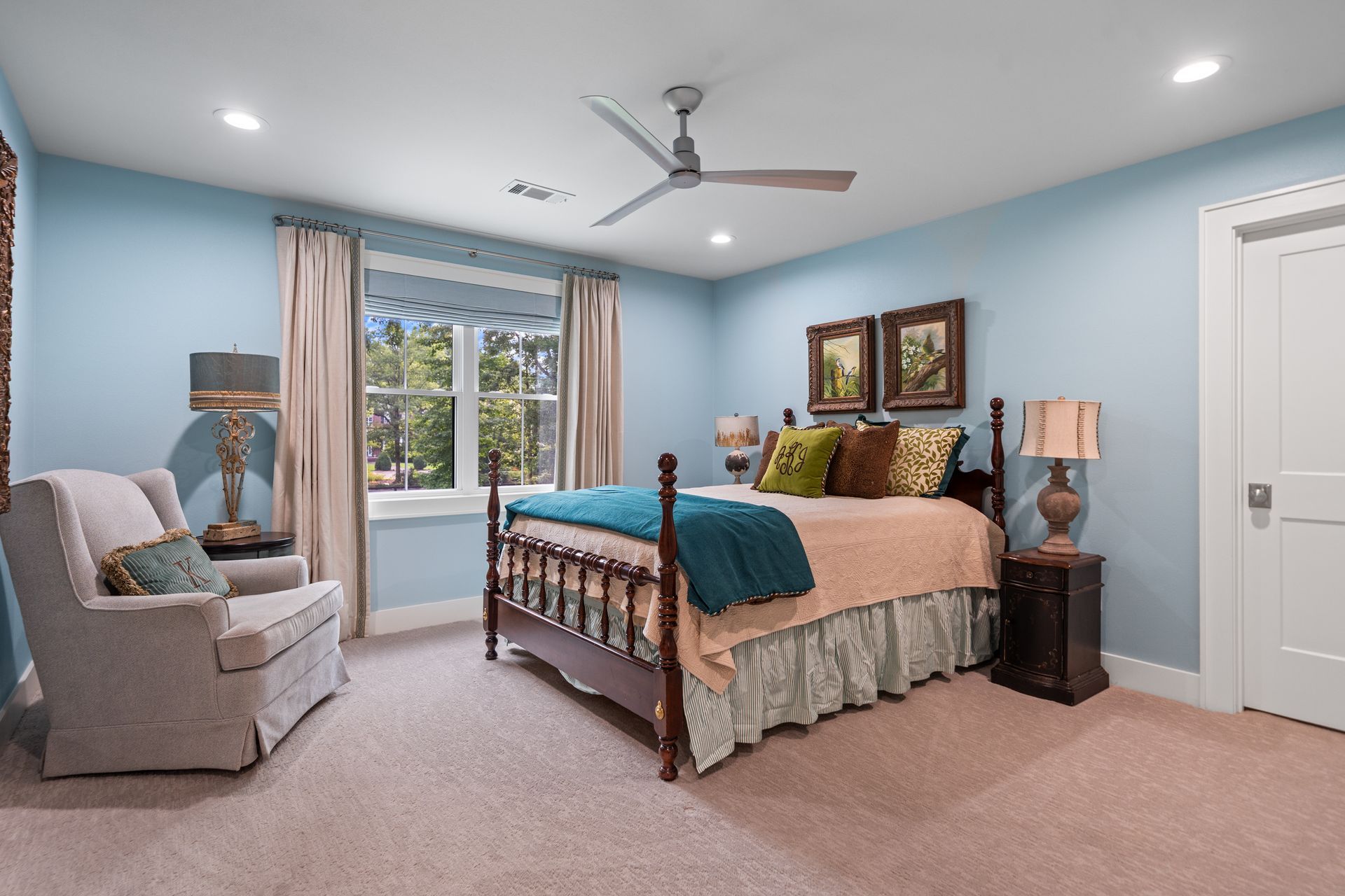 A light blue bedroom with a dark wood bed, a beige armchair, carpeted flooring, a ceiling fan, and a window with curtains.