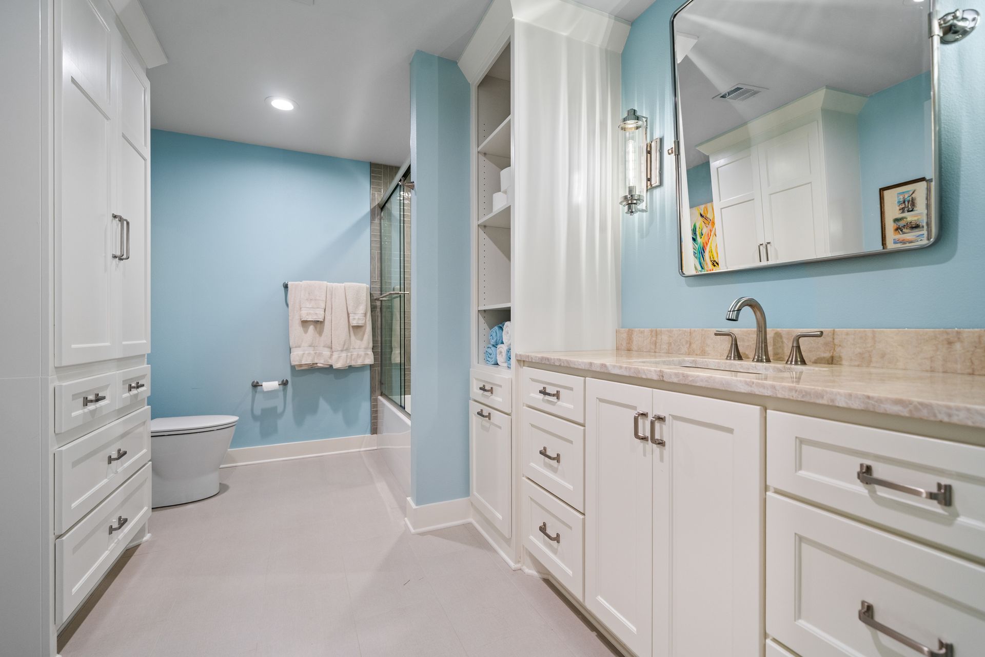 A bright bathroom with light blue walls, white cabinetry, a single sink, a mirror, and a walk-in shower.