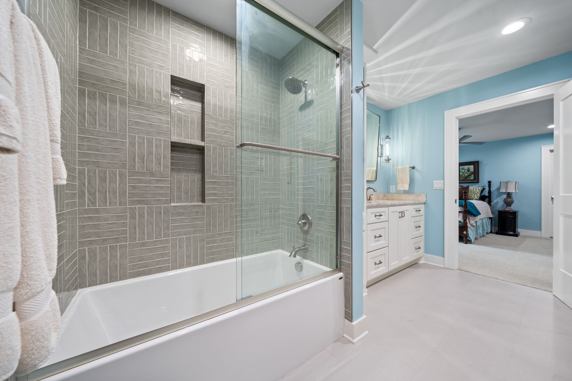 A bright bathroom with a white bathtub, gray patterned tile shower wall, glass door, and blue walls opening to a bedroom.