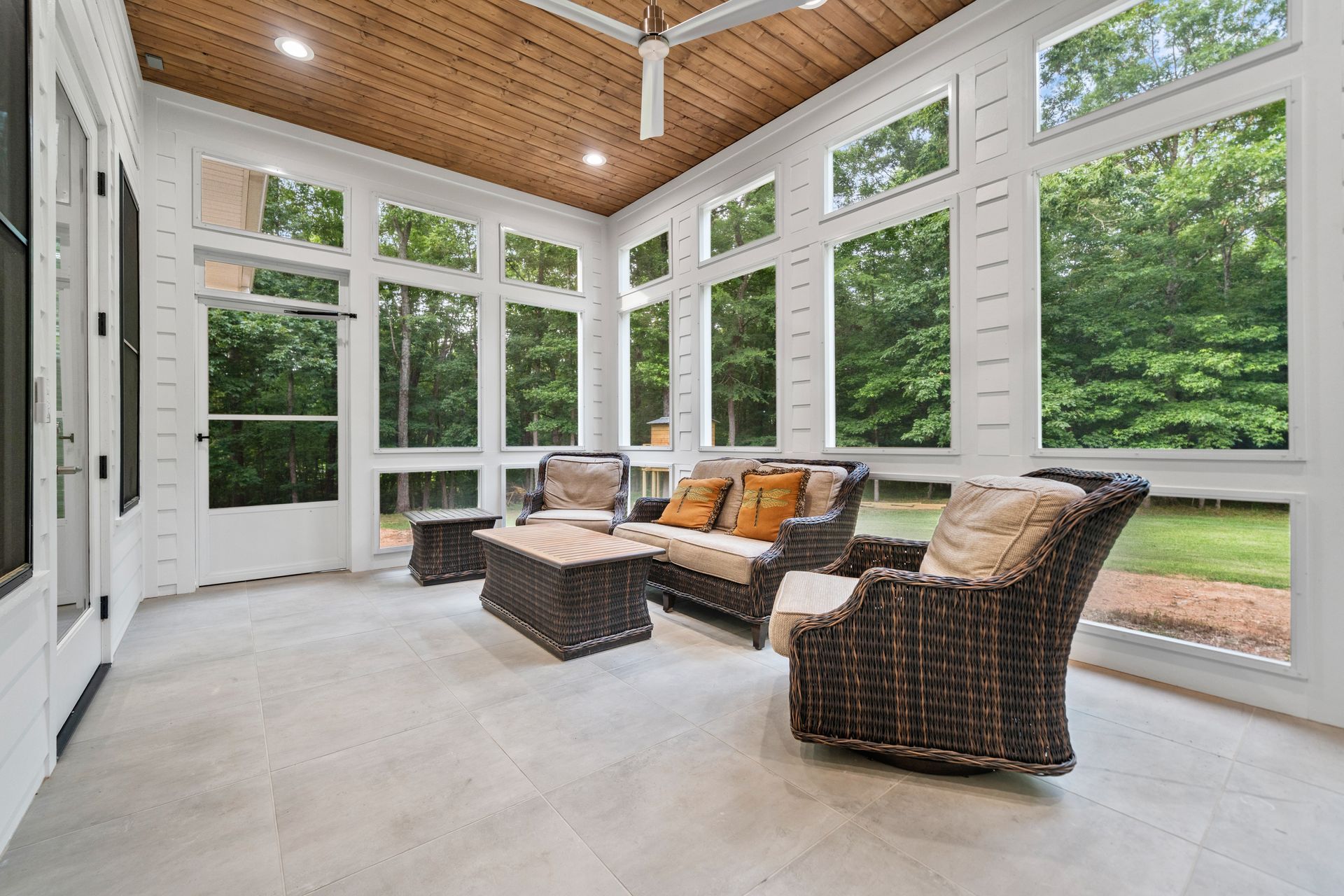 A bright sunroom with white walls, a wood-paneled ceiling, large windows, and wicker furniture on light tile flooring.