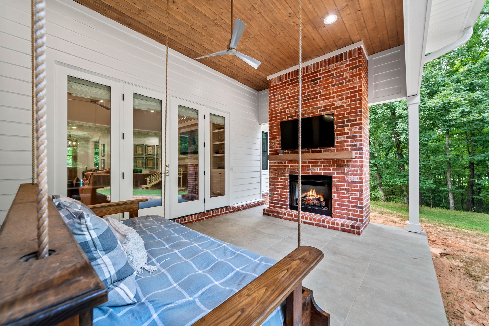 A covered patio featuring a hanging wooden swing, a brick fireplace with a mounted television, and views of a forest.