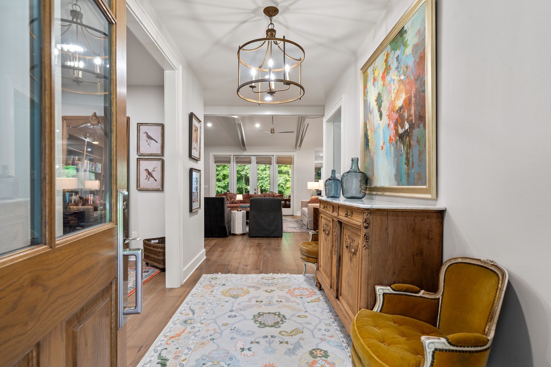A hallway with a wooden cabinet, mustard-yellow chair, large abstract painting, patterned rug, and lantern chandelier.