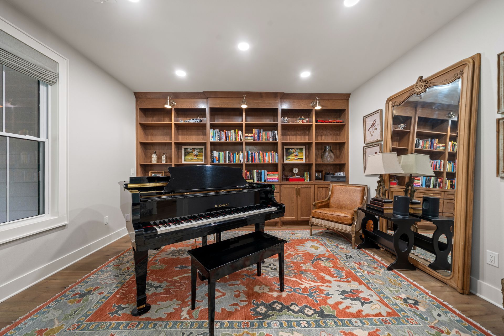 A grand piano, a leather chair, and a bookshelf in a room with a colorful patterned rug and a large floor mirror.