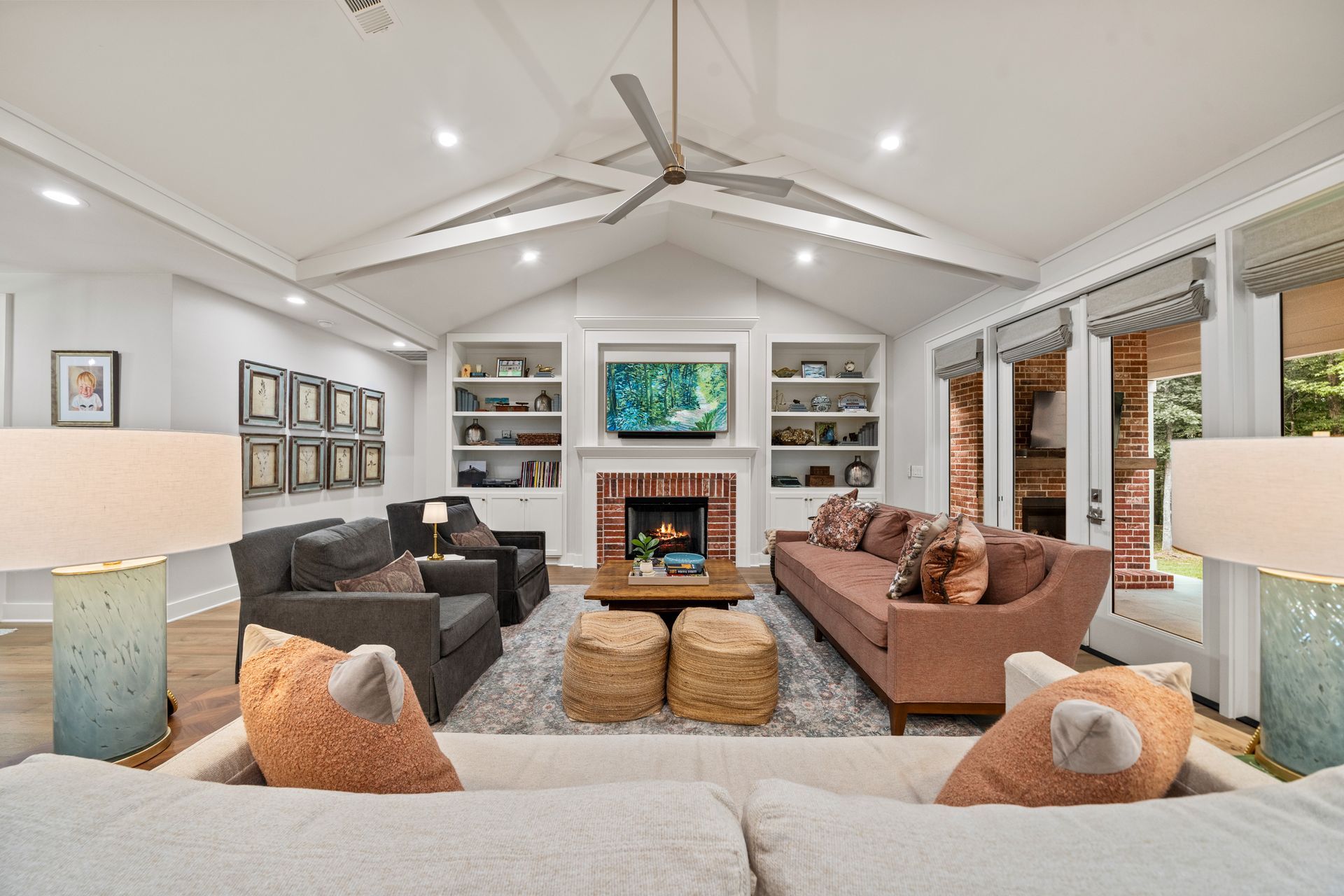 A living room with a vaulted ceiling, stone fireplace, bookshelves, a tan sofa, grey armchairs, and two textured lamps.