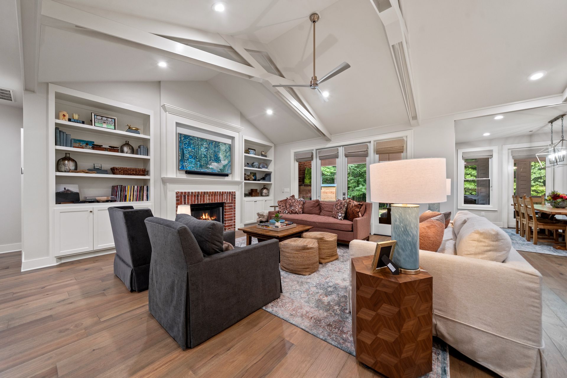 A bright, open-concept living room with a brick fireplace, built-in shelves, plush seating, and wood flooring.