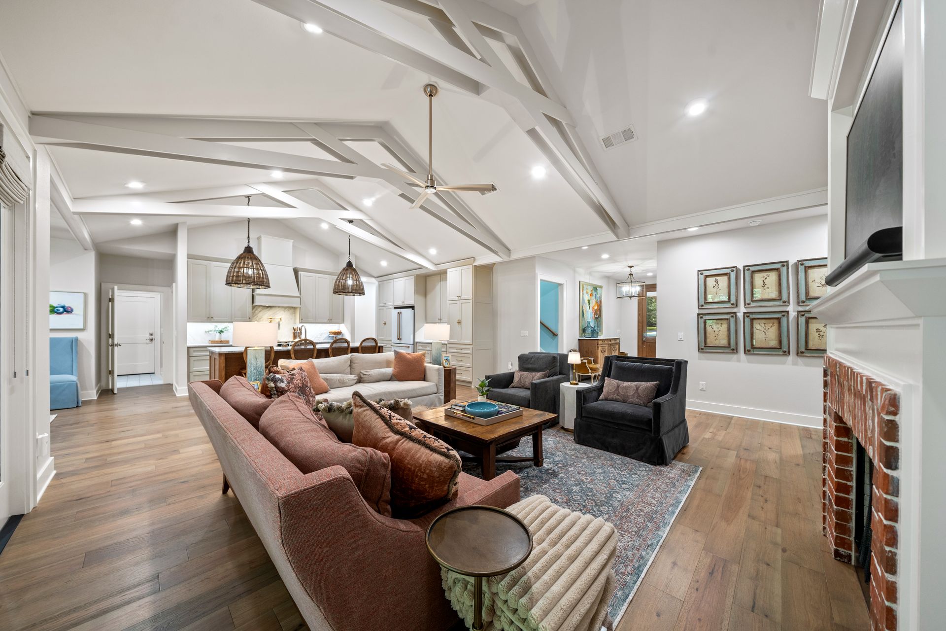 Open-concept living room with white vaulted ceilings, brick fireplace, grey couch, armchairs, and wood floors.