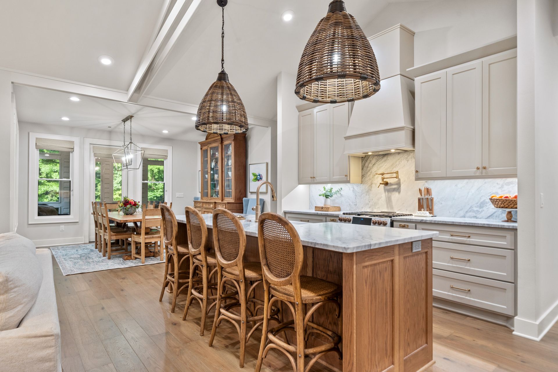 A bright, modern kitchen featuring a wood island with three wicker bar stools, pendant lights, and a dining area beyond.