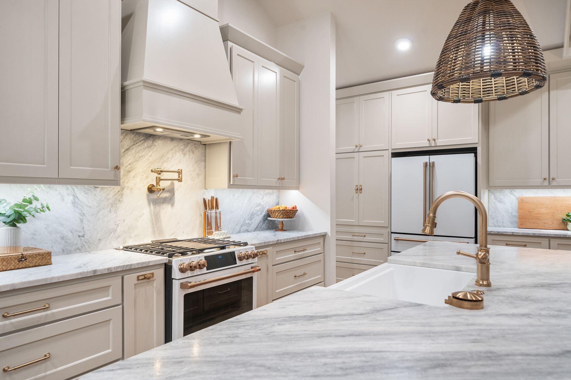 A modern kitchen features light-gray cabinetry, white marble countertops, a gold faucet, and a large woven pendant light.