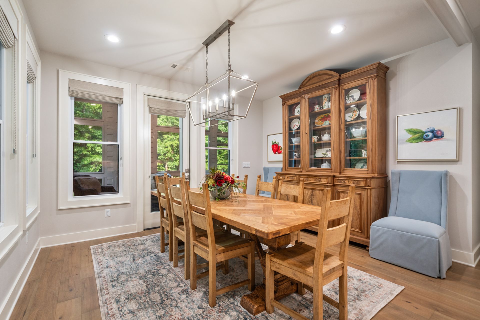 A wooden dining table and chairs set under a light fixture, with a matching hutch and a blue chair in a light-filled room.