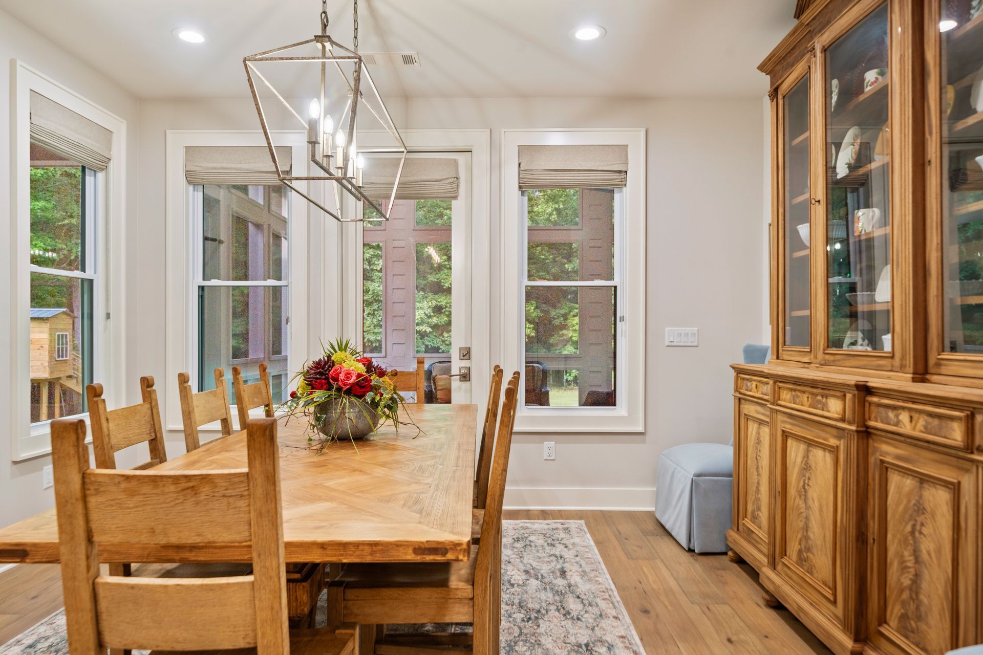 A dining room with a wooden table, chairs, a decorative chandelier, windows, and a large wooden hutch against a wall.