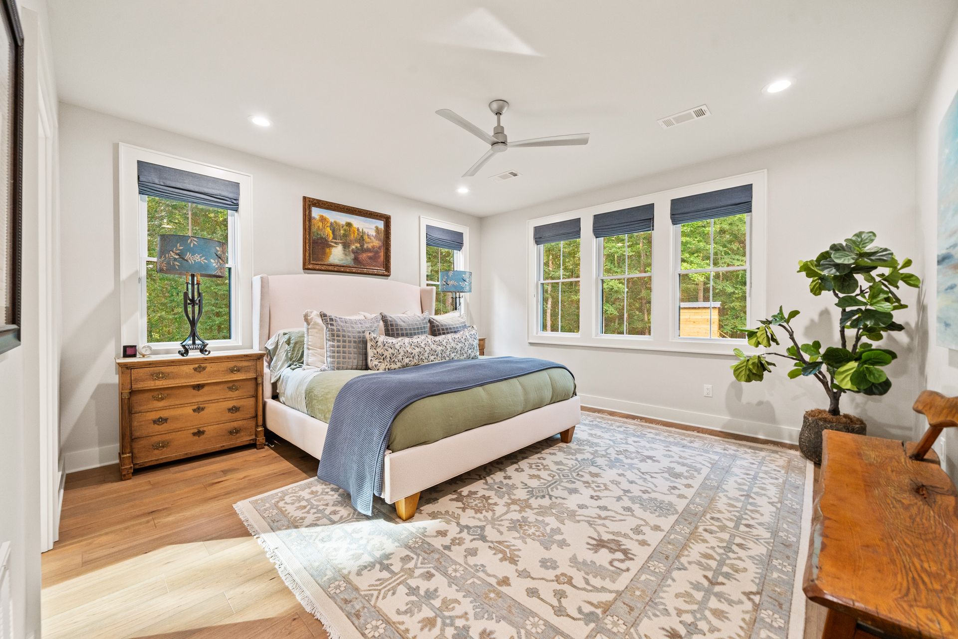 A bright, neutral-toned bedroom with a light upholstered bed, wooden dresser, large patterned rug, and potted plant.