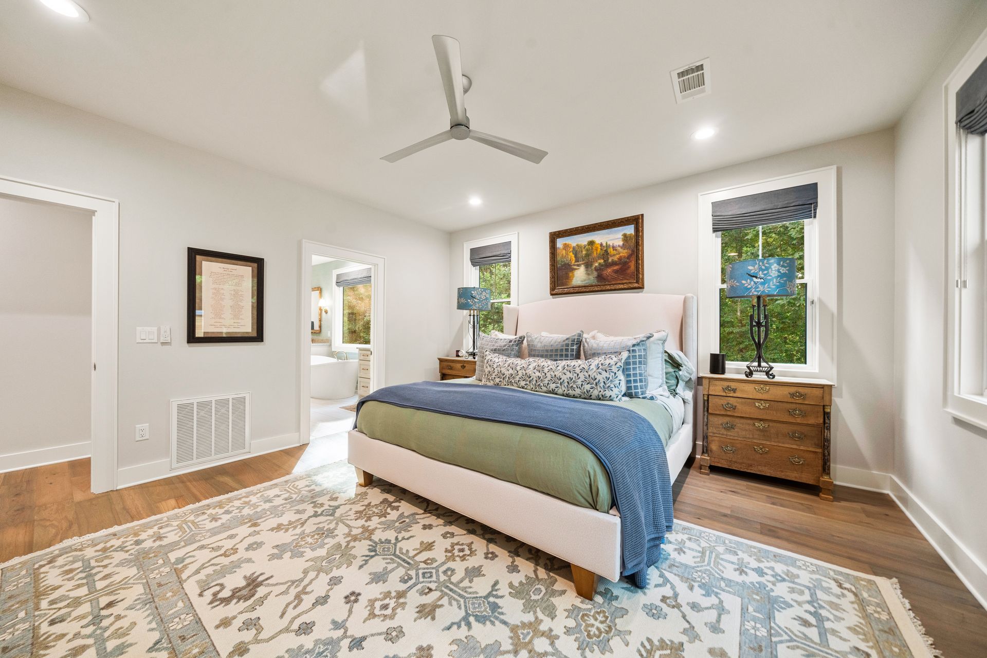 Bright bedroom with a king bed, patterned rug, wooden nightstand, ceiling fan, and white walls.