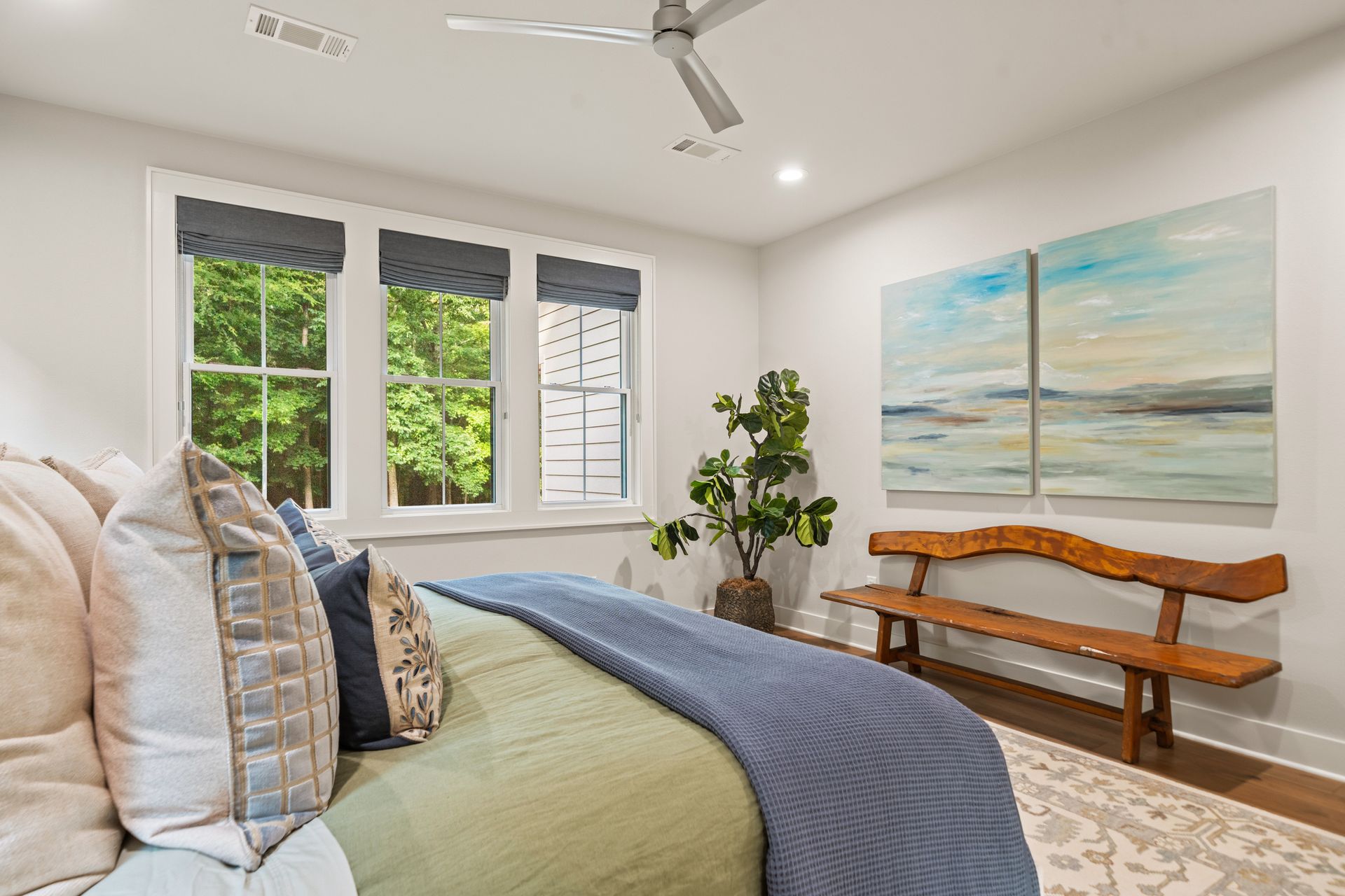 A bedroom featuring a bed with blue and green bedding, a rustic wooden bench, two large landscape paintings, and windows.