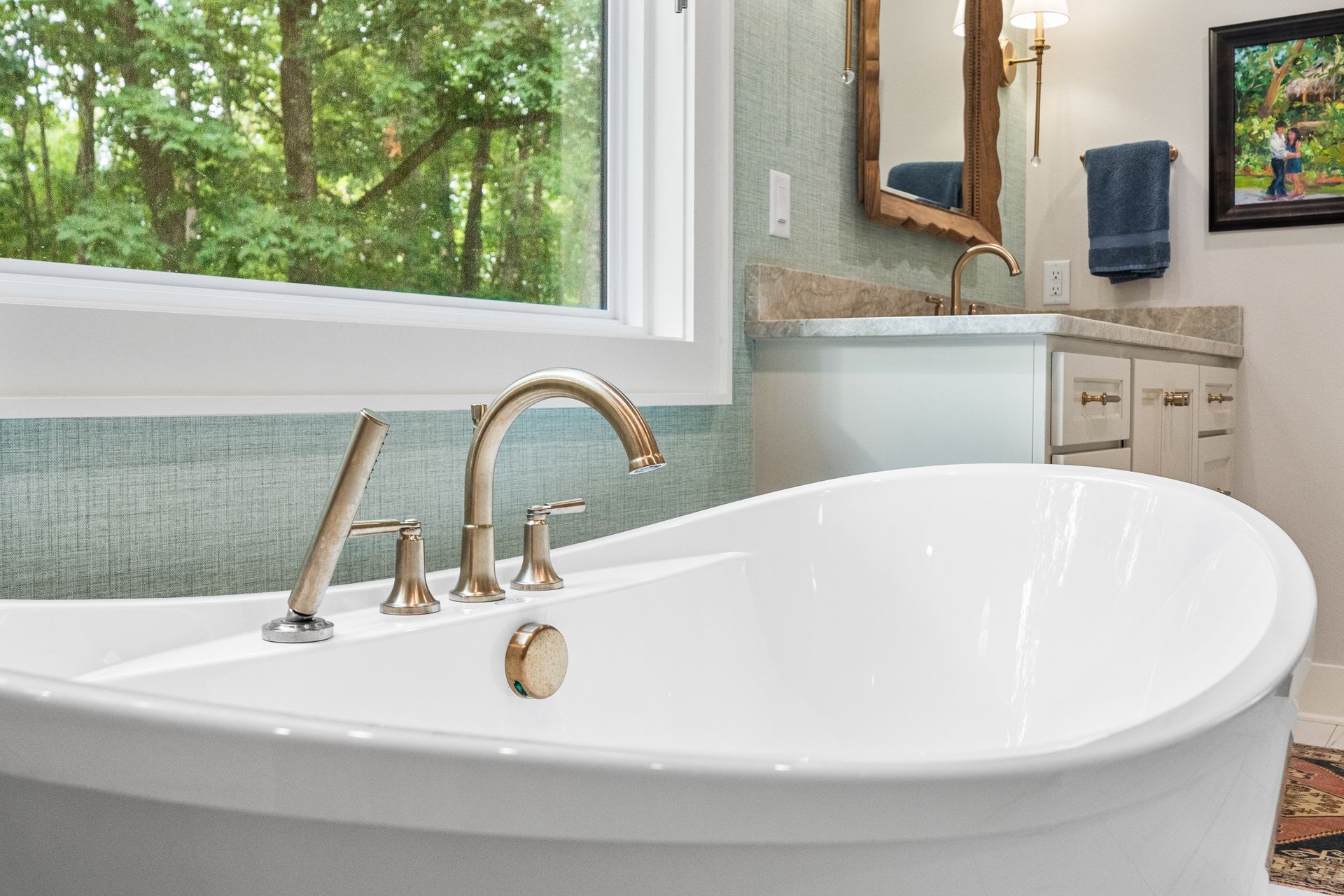 A white freestanding bathtub with brass fixtures sits in front of a window overlooking green trees in a modern bathroom.