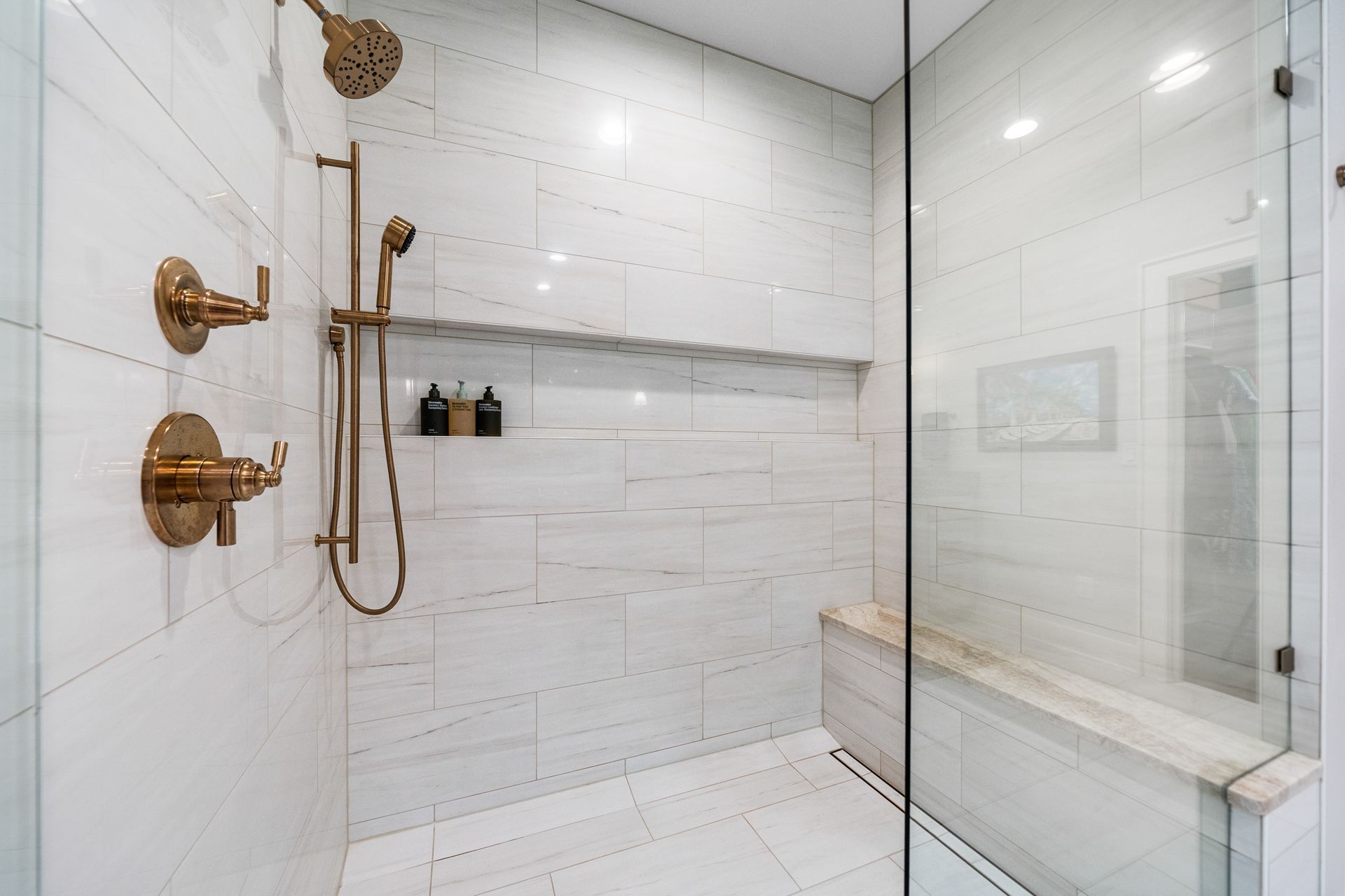 A modern walk-in shower with white marbled tile walls, brass fixtures, a handheld showerhead, and a tiled built-in bench.