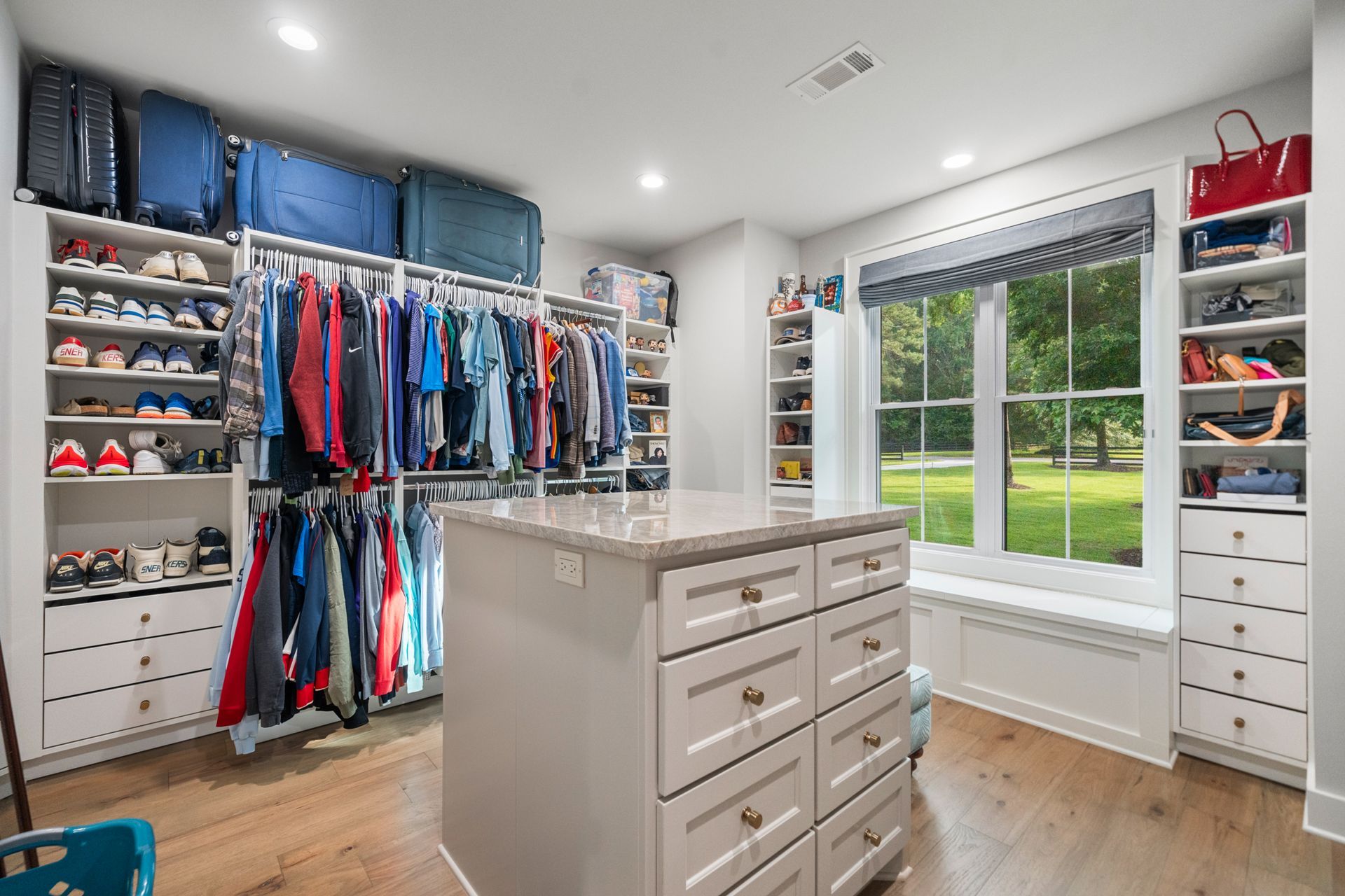 A spacious, well-organized walk-in closet with hanging clothes, shelving, a central storage island, and a window seat.