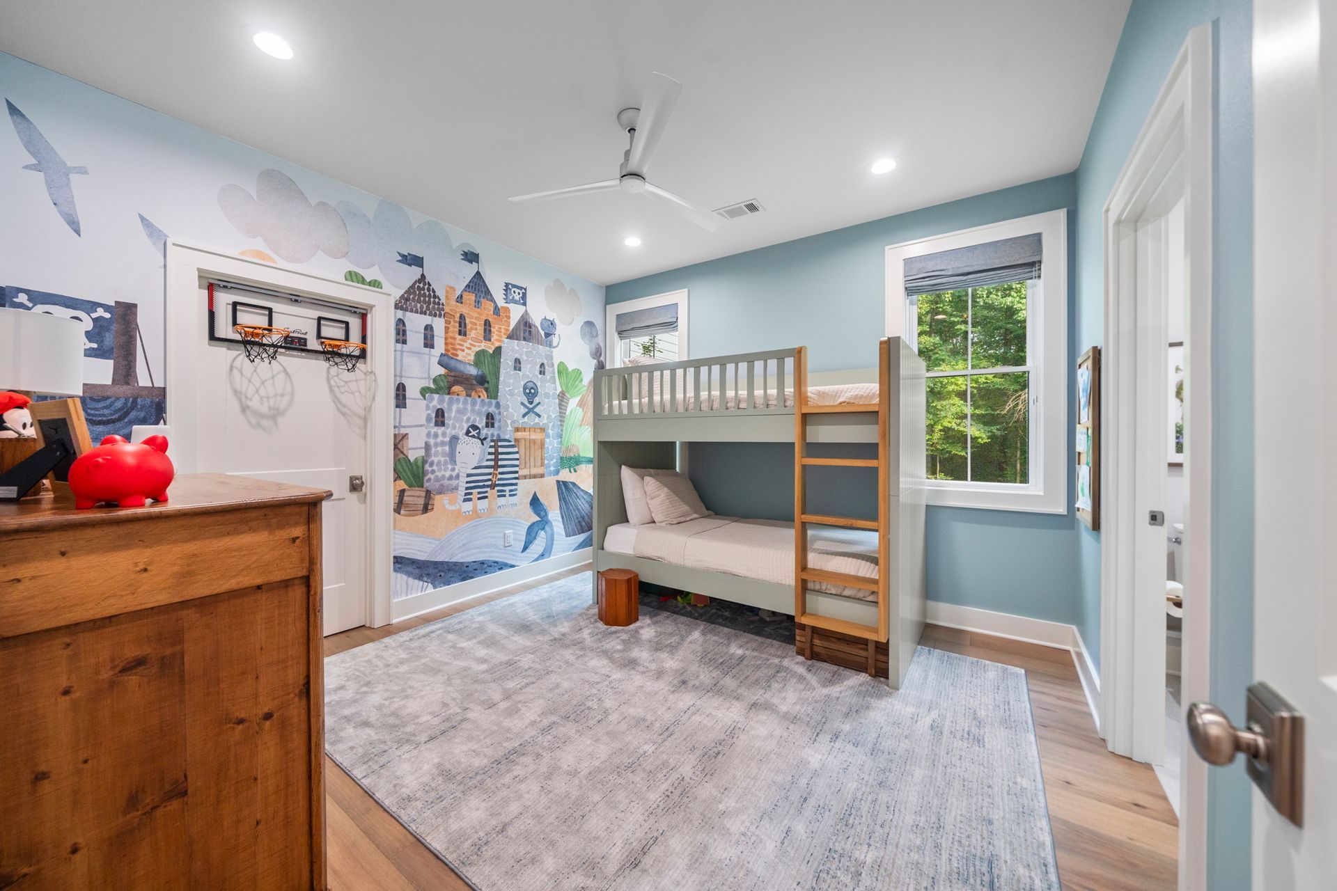 A children’s bedroom features a grey bunk bed, a castle wall mural, a light blue rug, and a rustic wooden dresser.