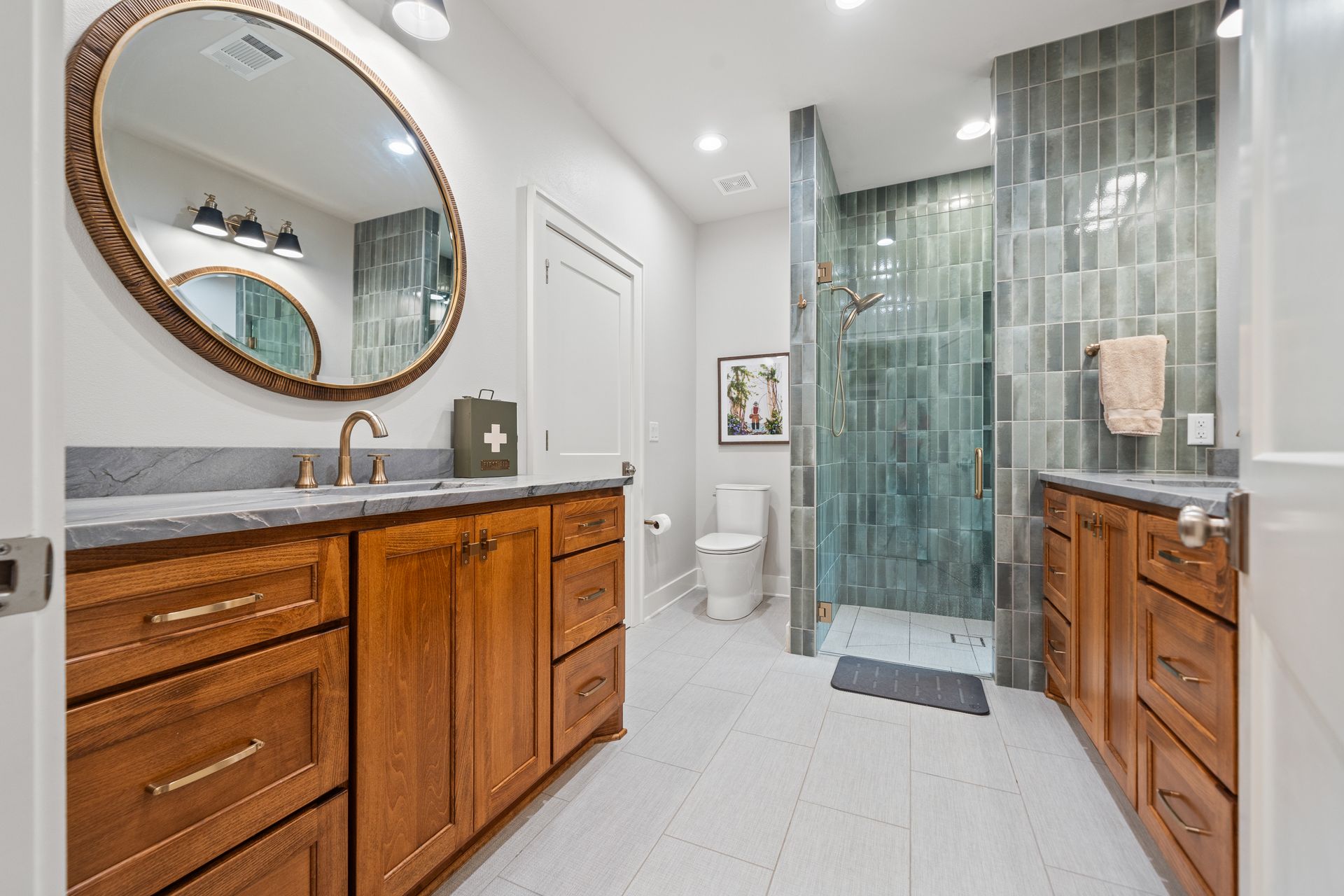 Modern bathroom with wood vanities, oval mirror, gray tile walk-in shower, and white tile flooring.
