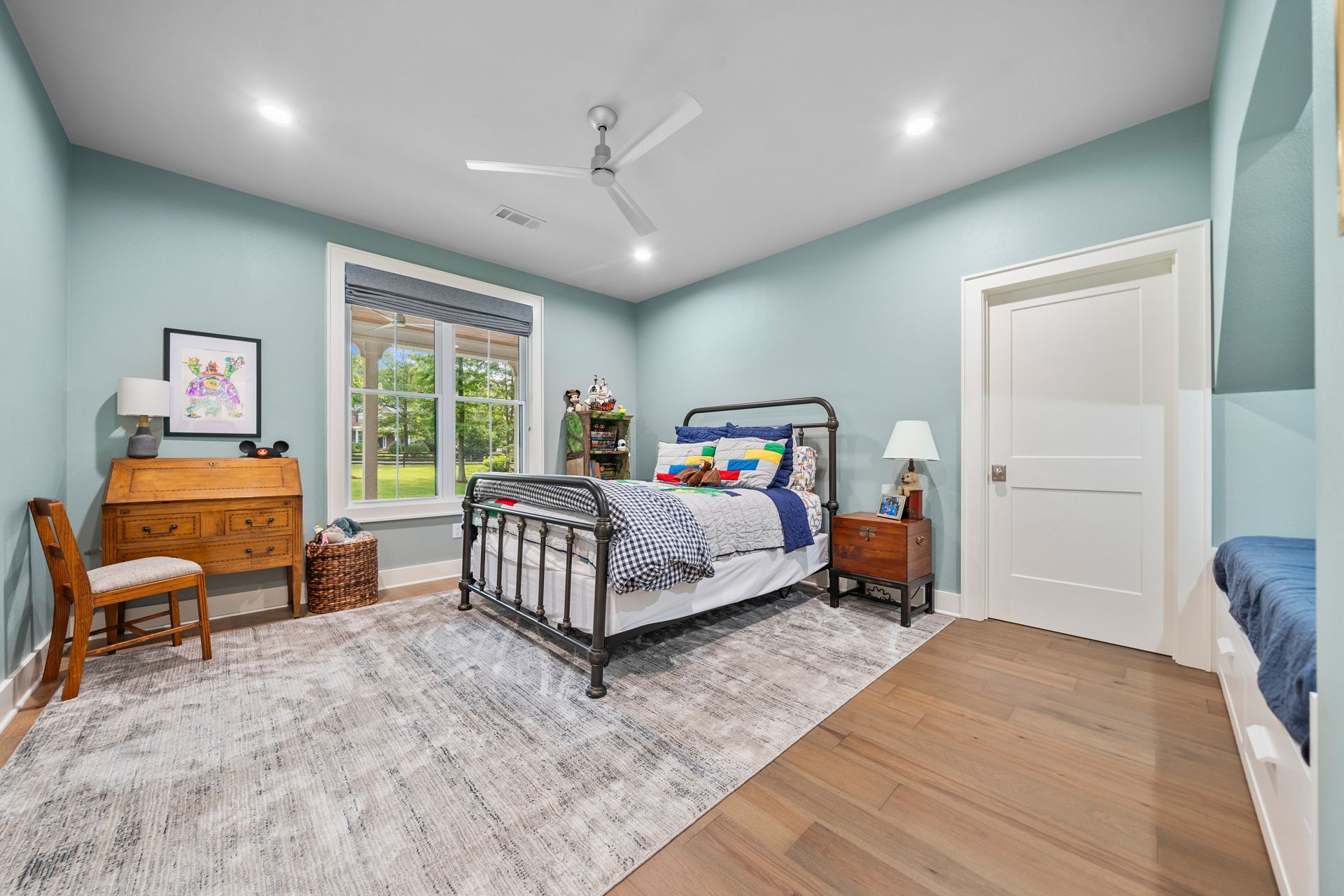 A bedroom with light blue walls, a metal bed frame, wooden desk, area rug, and ceiling fan.