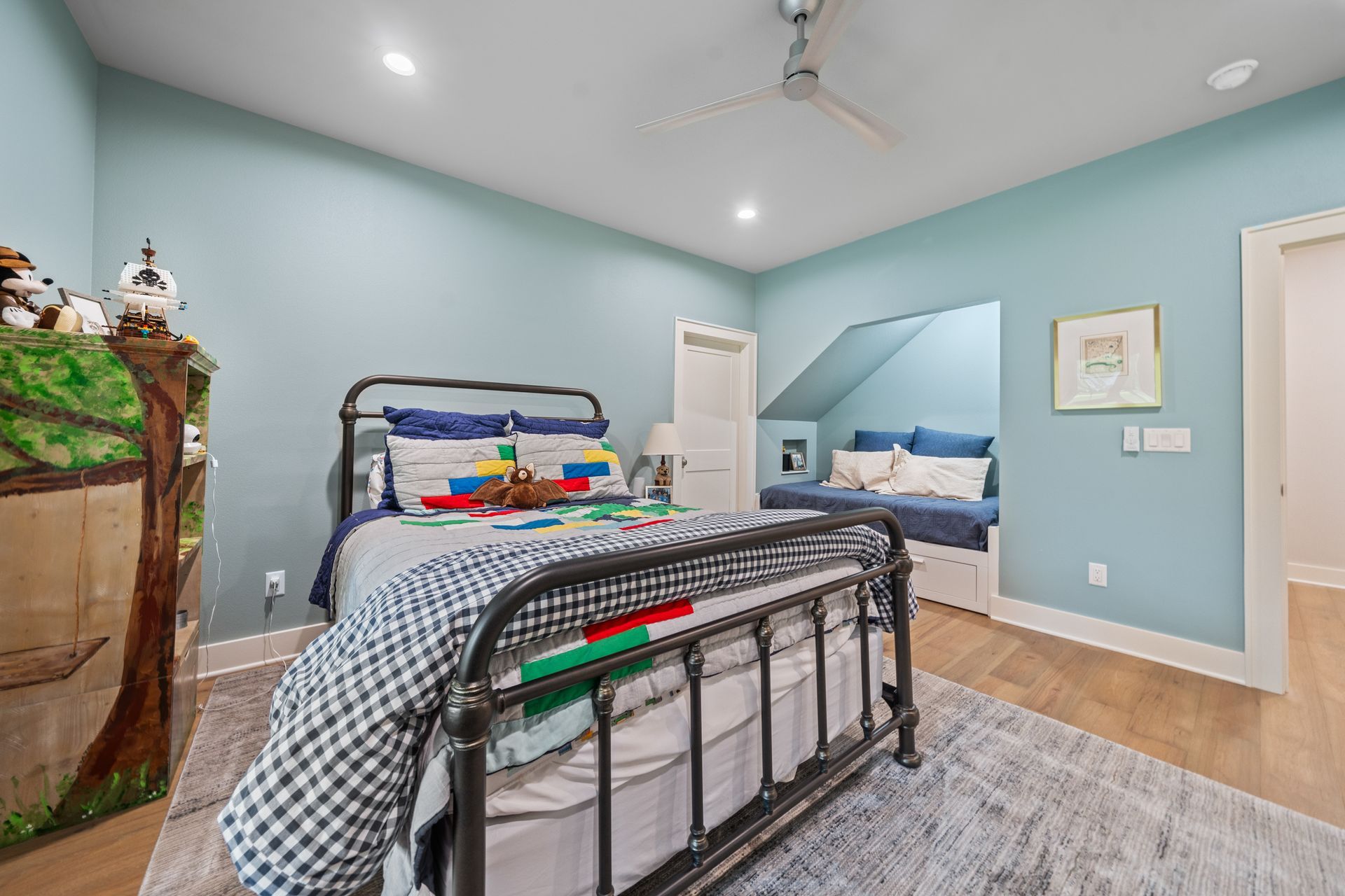 A bedroom with light blue walls, a metal-framed bed, a built-in reading nook, and a rug on hardwood floors.