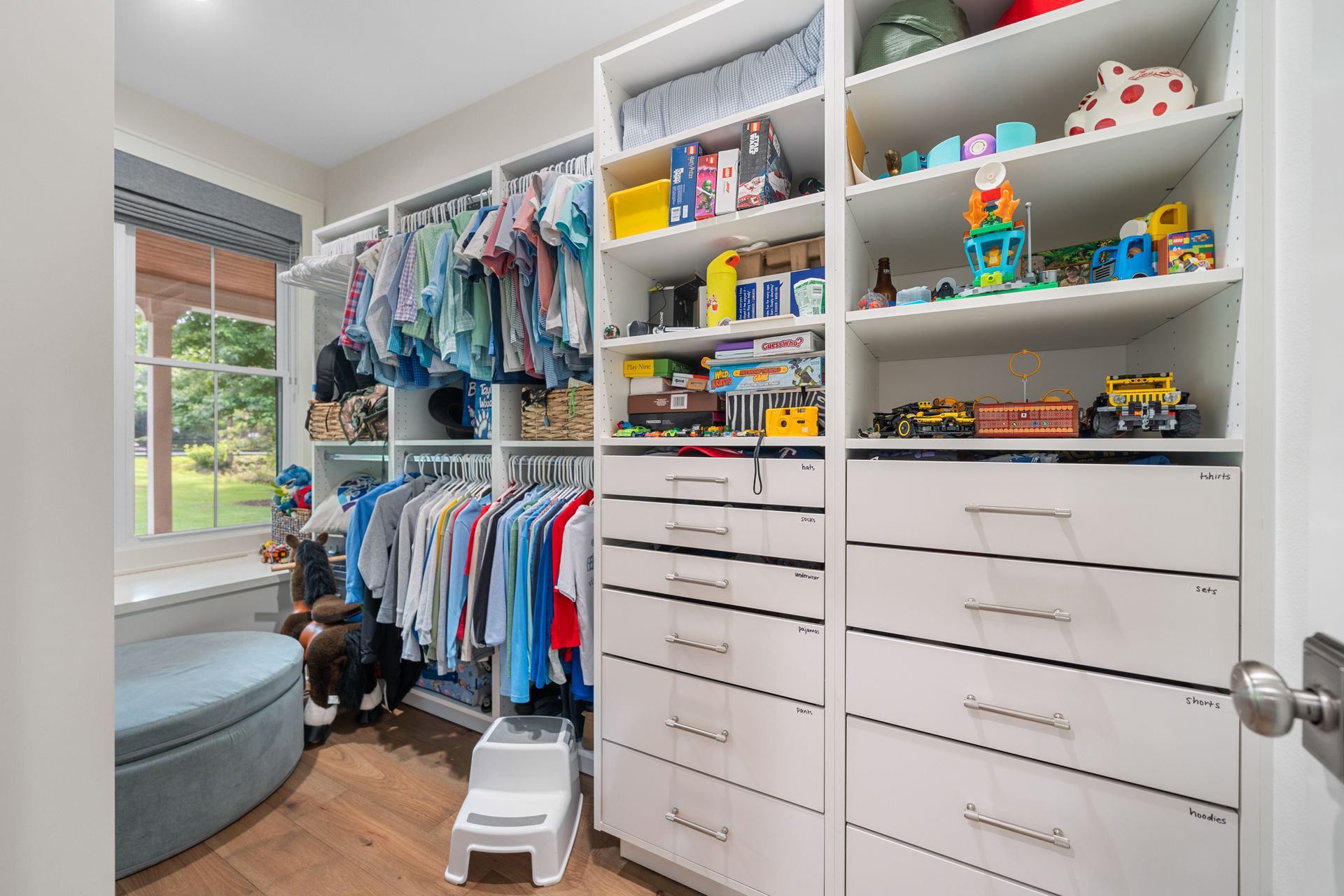 A brightly lit walk-in closet featuring organized hanging clothes, shelves with toys, and white drawer units.