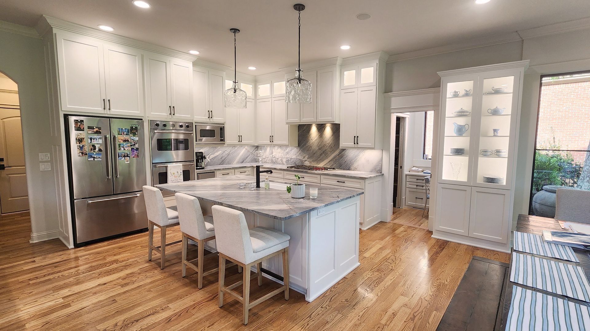 A modern white kitchen featuring a marble-topped island with three white stools, stainless appliances, and wood flooring.