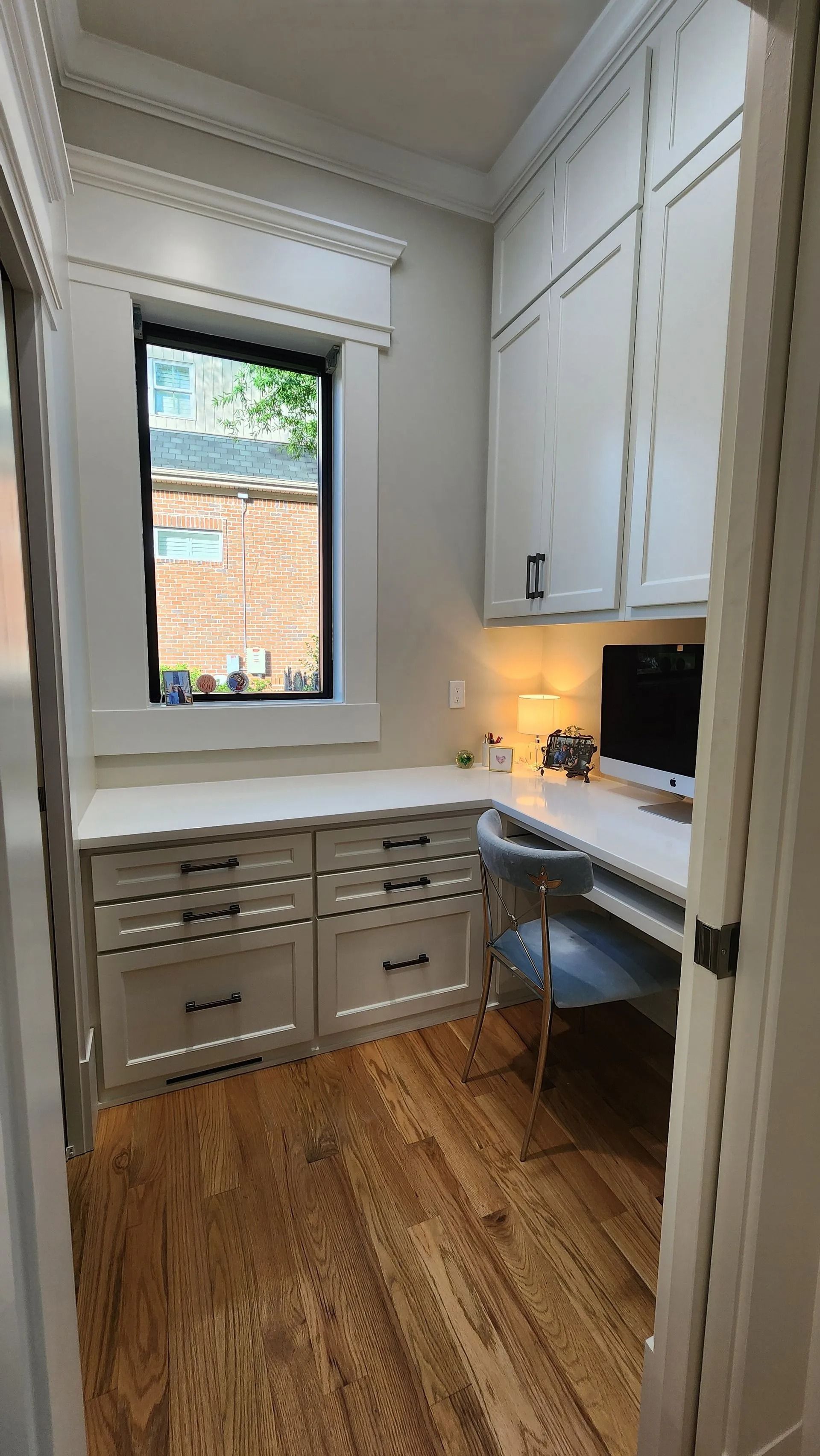 A small, bright home office with white cabinetry, a desk, a computer, and hardwood floors next to a window.