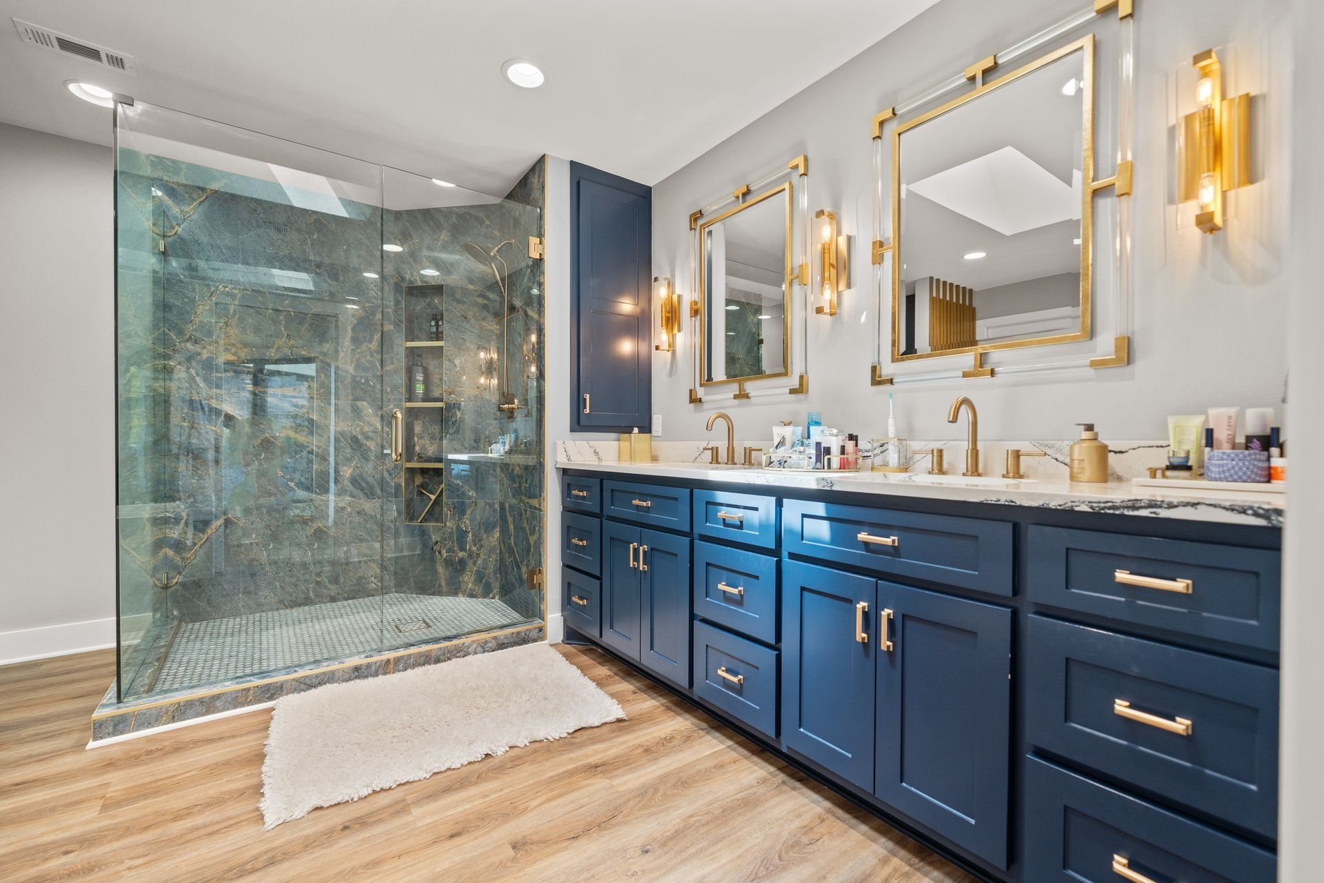 A modern bathroom with a glass-enclosed shower, a long navy blue vanity with gold hardware, and two gold-framed mirrors.